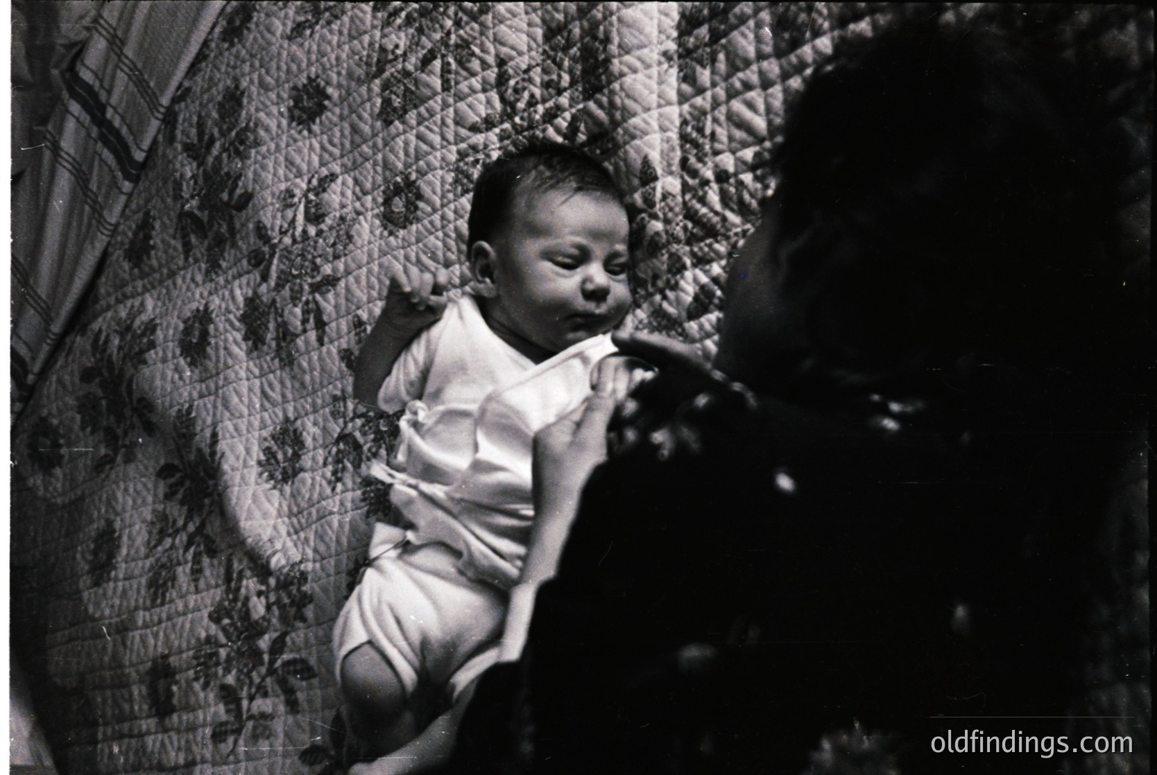 Black-and-white portrait of an infant in a hammock-style bed, draped in a woven, textured fabric. The child wears a simple short-sleeve shirt and diaper. Likely mid-20th century domestic setting, emphasizing simplicity and warmth.
