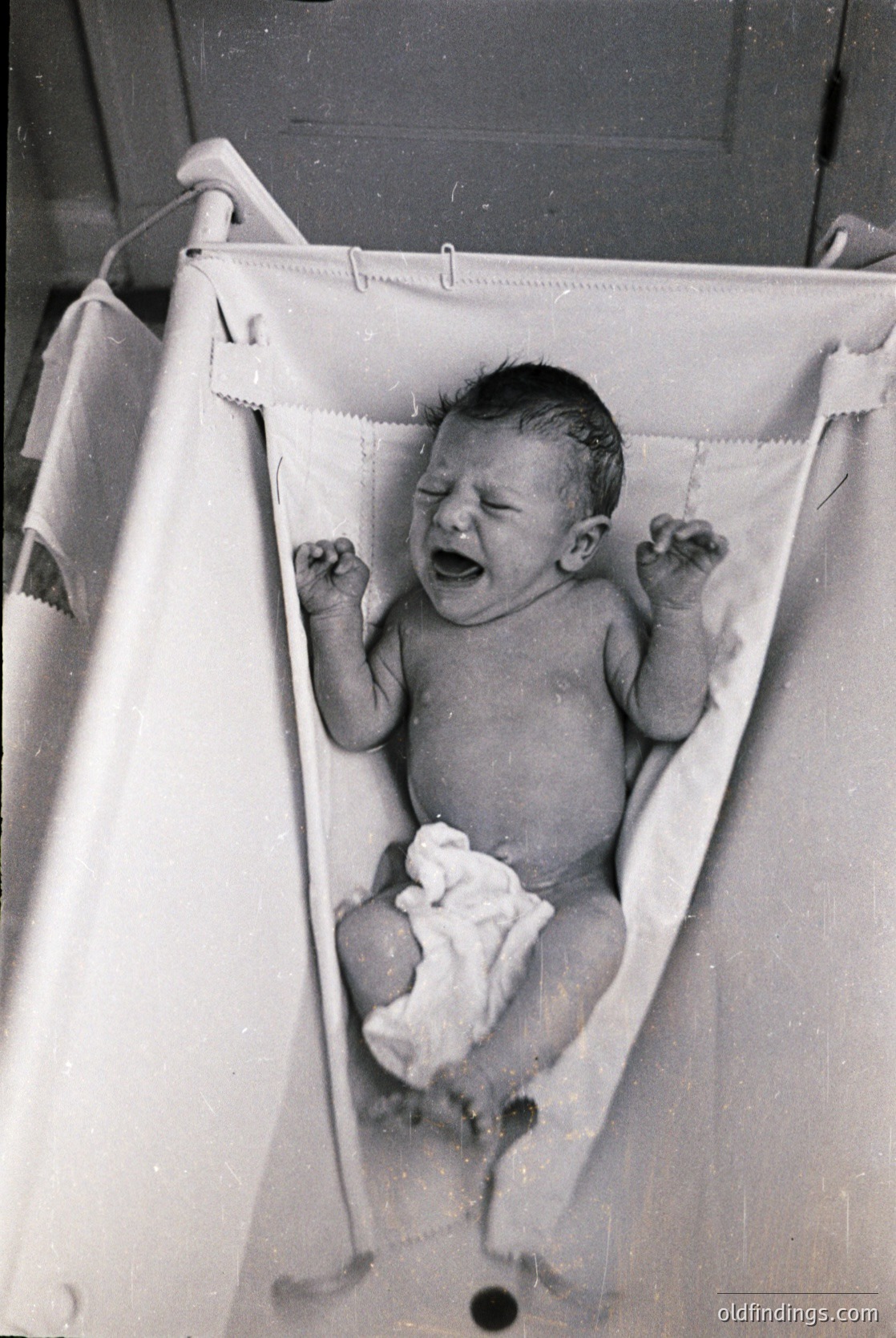 Vintage black-and-white photo of an infant in a cloth baby bath sling, mid-cry with arms raised. Soft towel draped over shoulders. Likely mid-20th century domestic setting.