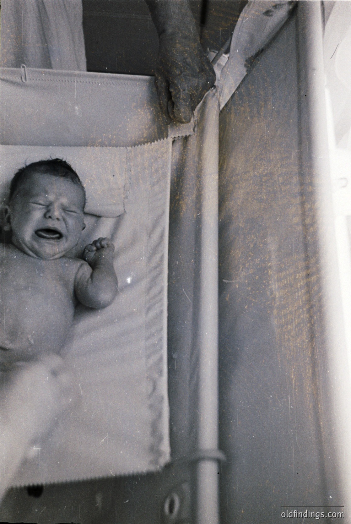 Vintage black-and-white photo of an infant in a metal-framed bassinet, crying with hand near mouth. Soft fabric lining shows wear. Likely mid-20th century domestic setting.