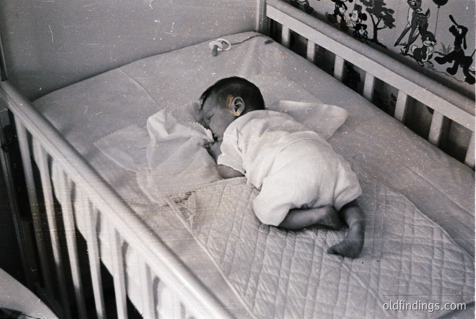 Vintage black-and-white photo of an infant sleeping in a metal-framed crib with floral wallpaper in background. Simple white onesie and diaper visible. Likely mid-20th century domestic setting.