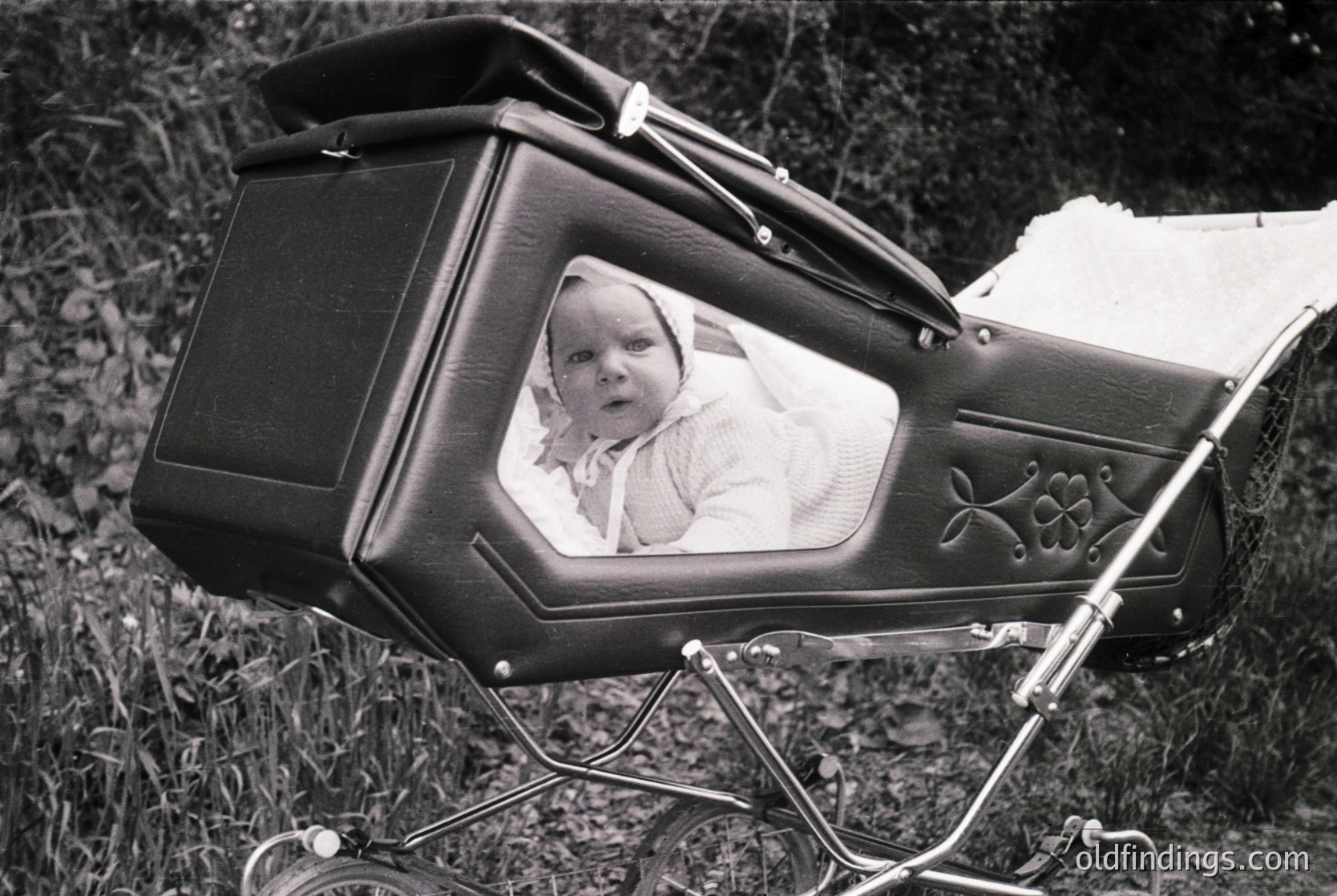 Vintage black-and-white photo of a child in a classic wicker pram with floral embroidery, set outdoors on grass. The pram’s curved canopy and metal detailing suggest mid-20th century design ( ).
