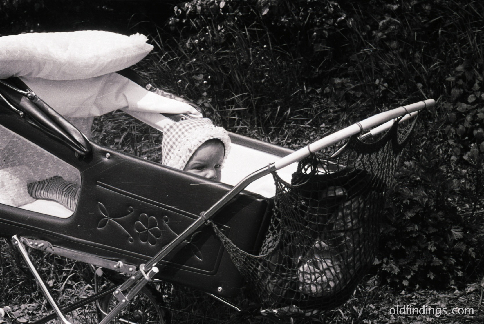 Vintage black-and-white photo of a child in a vintage pram with ornate metalwork, resting on grass. A fishing net and rod lie beside, suggesting outdoor leisure. Mid-20th century family lifestyle.