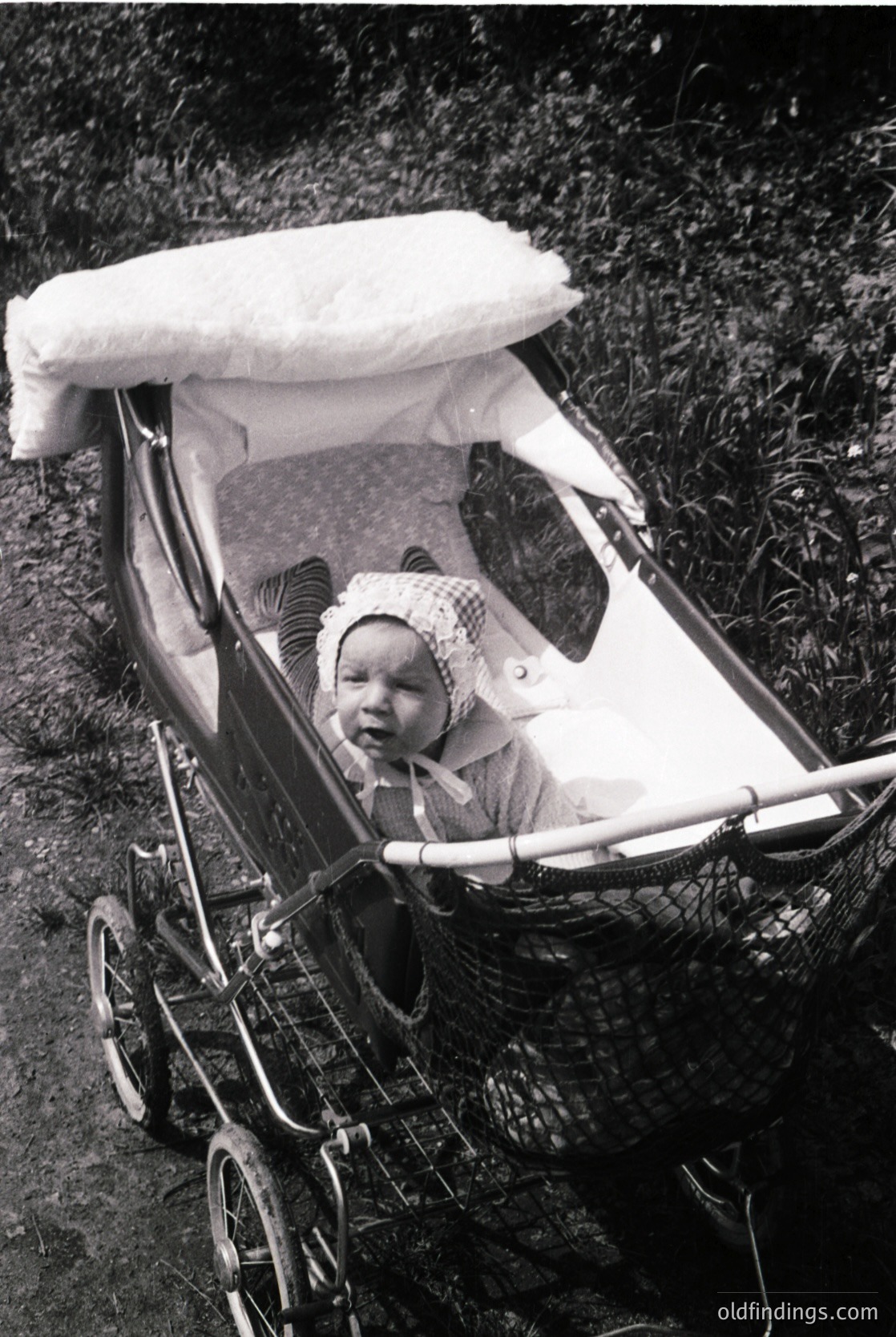 Vintage black-and-white photo of an infant seated in a classic metal-framed pram with a white canopy, set in an outdoor grassy area. The child wears a knitted hat and light-colored outfit, suggesting mid-20th century (1940s–1960s) fashion.