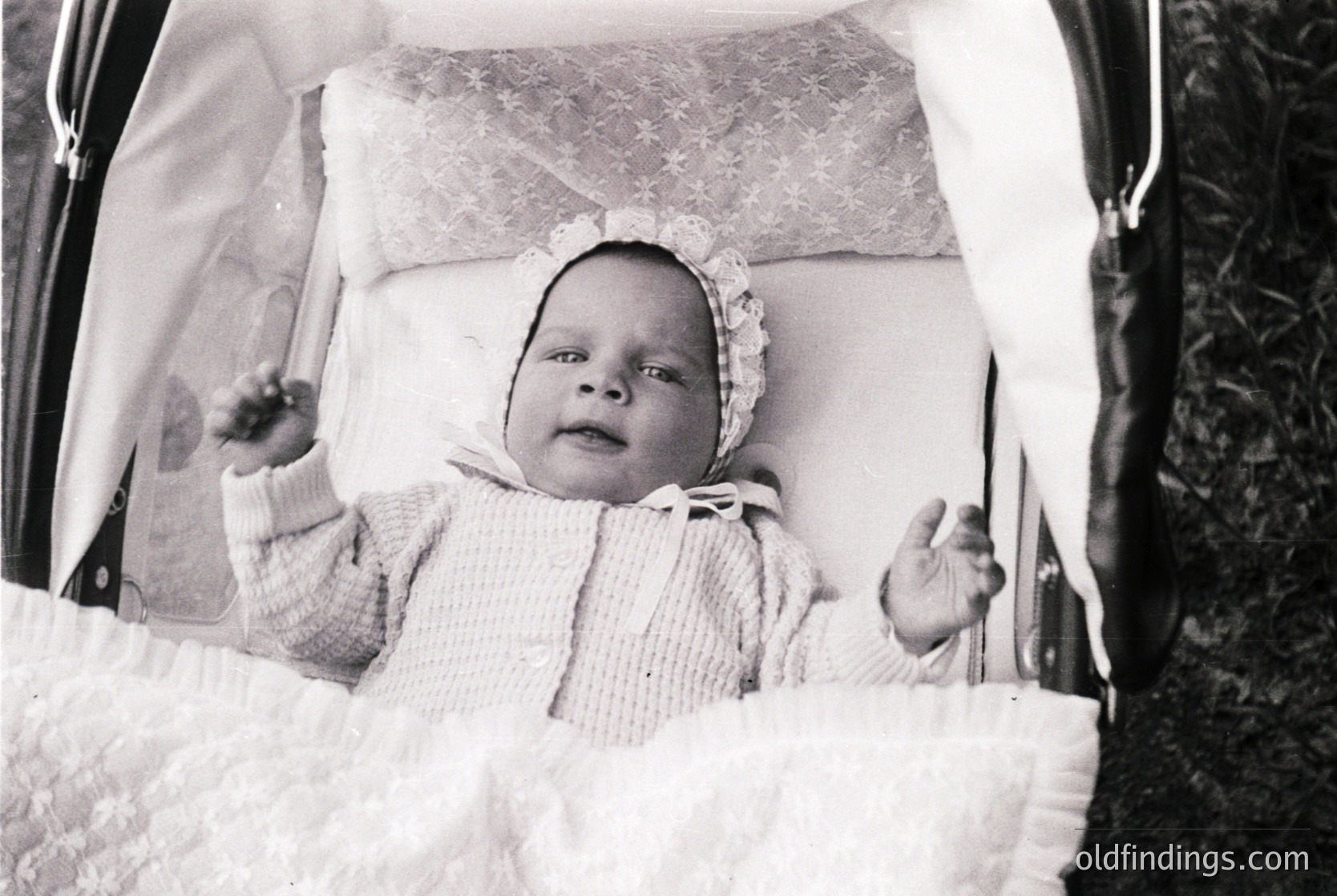 Vintage black-and-white photo of an infant seated in a vintage stroller, dressed in a knitted onesie and bonnet. Soft focus and outdoor setting suggest mid-20th century.