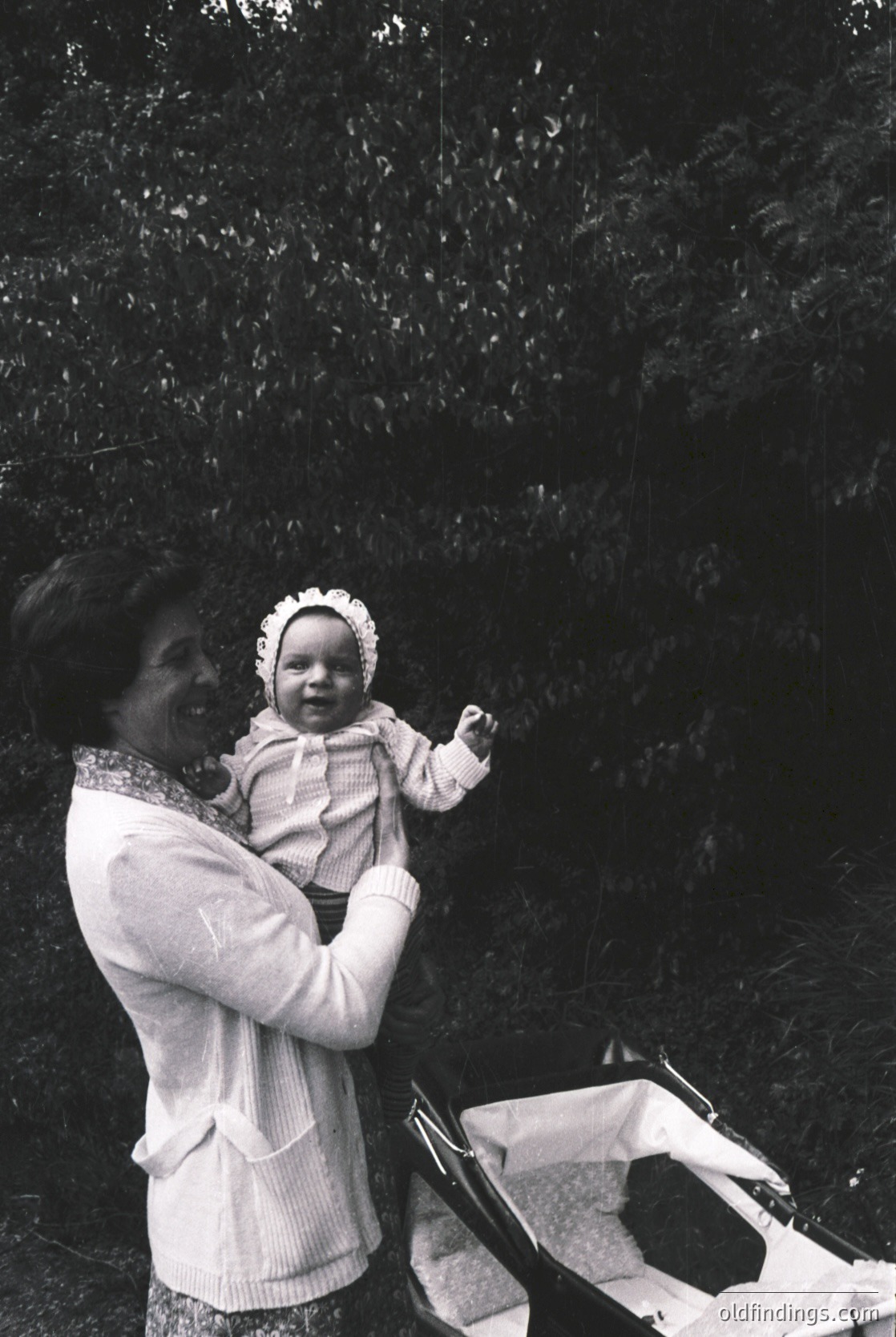 A woman in a vintage sweater and patterned scarf holds a baby in a knitted outfit with a ruffled hat, outdoors near dense foliage. A woven basket rests on the ground beside them, suggesting a picnic or nature outing. Style hints at mid-20th century (1950s–1960s).