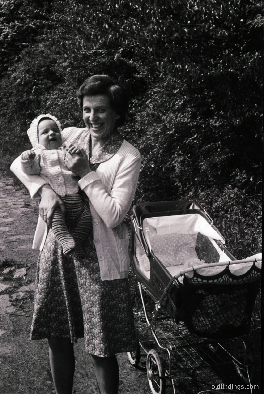 A woman in 1960s-era patterned dress and cardigan holds a baby in a striped onesie, posing outdoors near a vintage pram. Lush greenery and rocky terrain frame the scene.