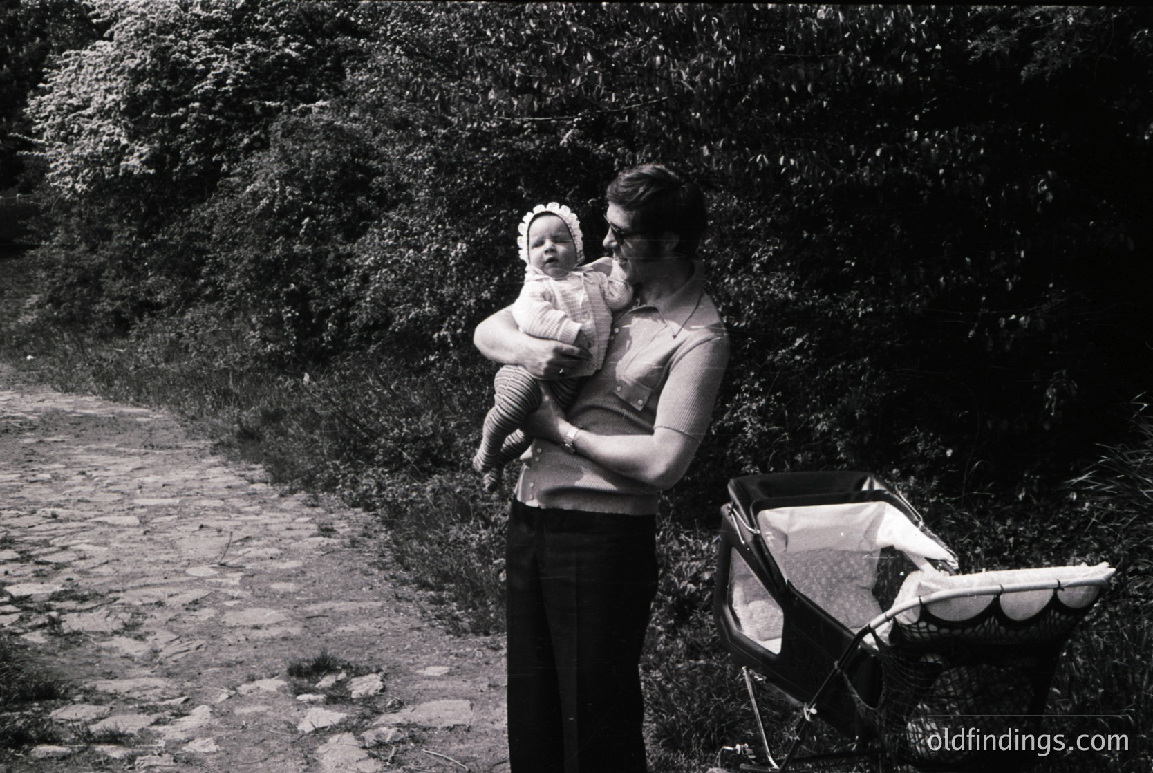 A woman cradles an infant in a rural pathway flanked by dense greenery, holding a folded stroller beside her. Mid-20th century attire and monochrome tone suggest or European countryside.