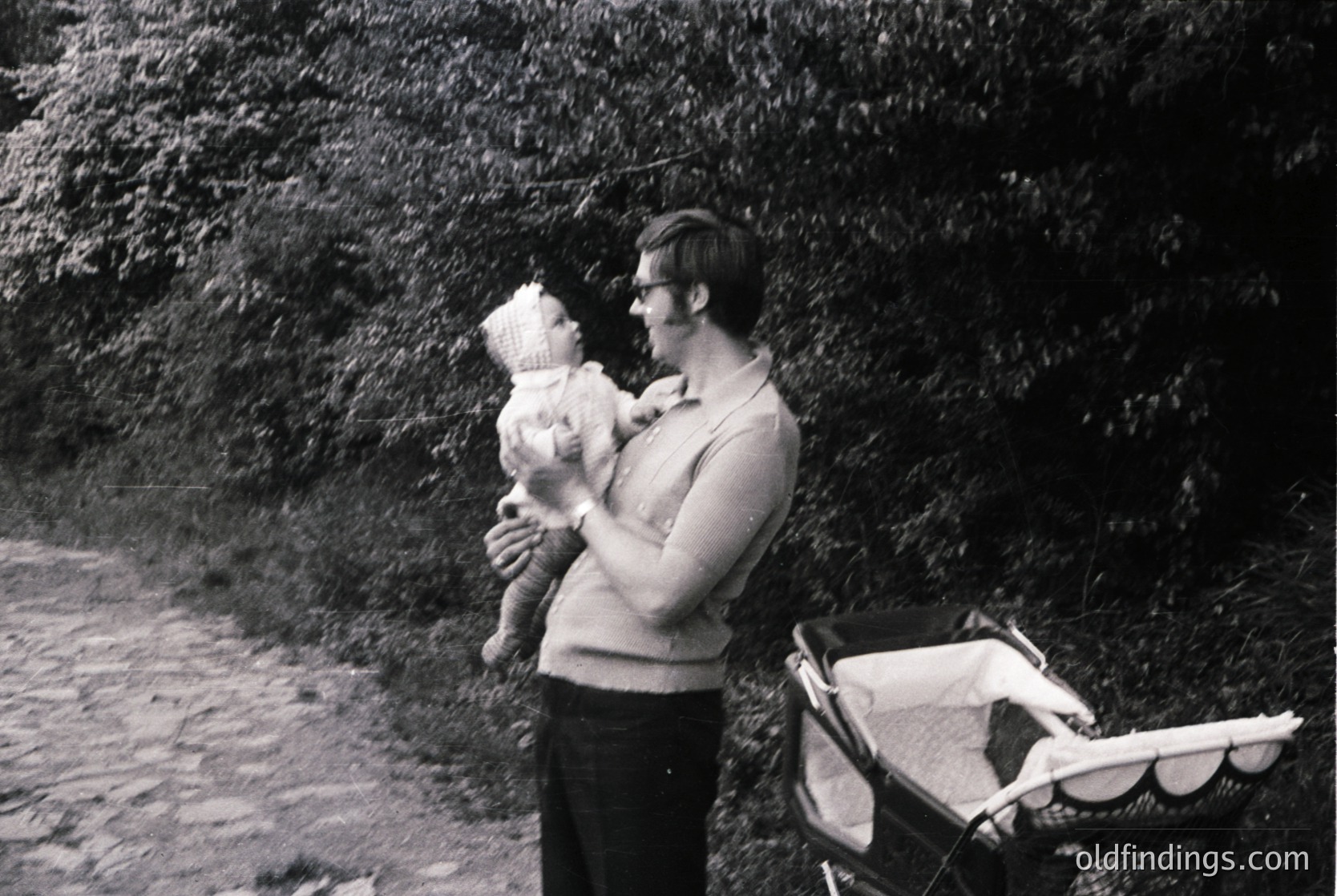 Mid-20th century black-and-white photo of a man in a sweater and glasses holding a baby outdoors, with a vintage pram in the background. Rugged terrain suggests a coastal or park setting.