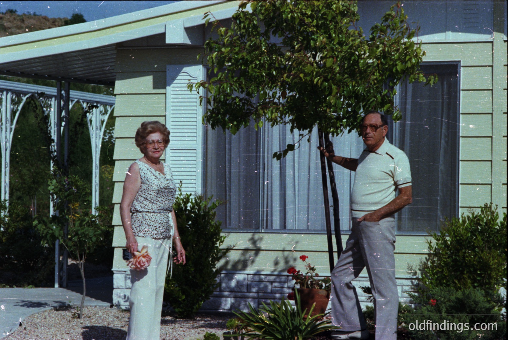 Mid-century modern home exterior with curved roof and sliding glass doors, 1960s USA. Elderly couple in retro attire—woman in floral dress, man in short-sleeved polo and khakis—pose outdoors. Lush greenery and manicured garden with potted plants.