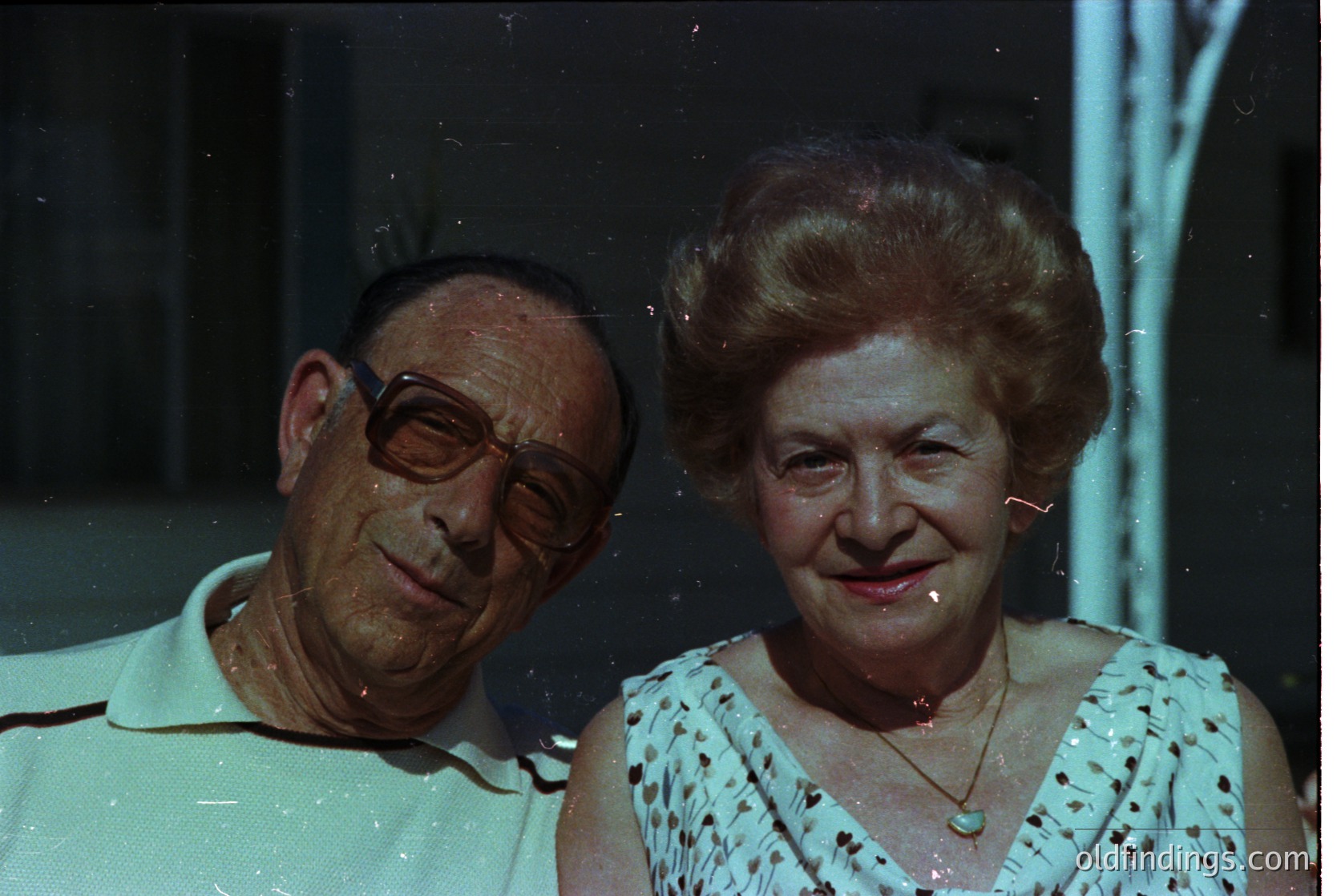 Vintage portrait of an elderly couple posing indoors, likely mid-20th century. Man wears round glasses and a white polo shirt; woman has a voluminous updo and floral-patterned blouse. Soft focus and slight film grain suggest 1960s–1970s photography.