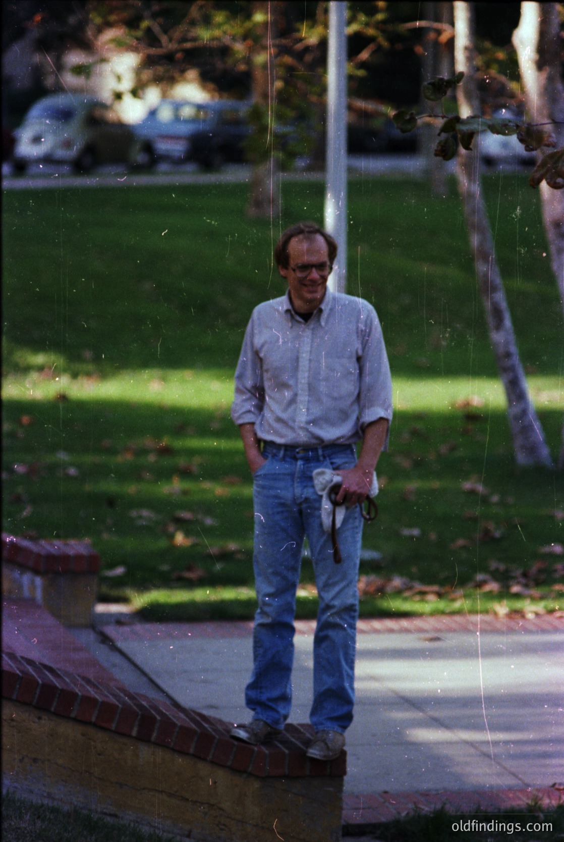 Mid-century man in a light blue button-down and rolled-up jeans stands on a brick patio, holding a cane. Reflections of trees and parked cars blur the background, suggesting an urban park or campus setting. Film grain and sepia tone indicate a vintage or photograph.