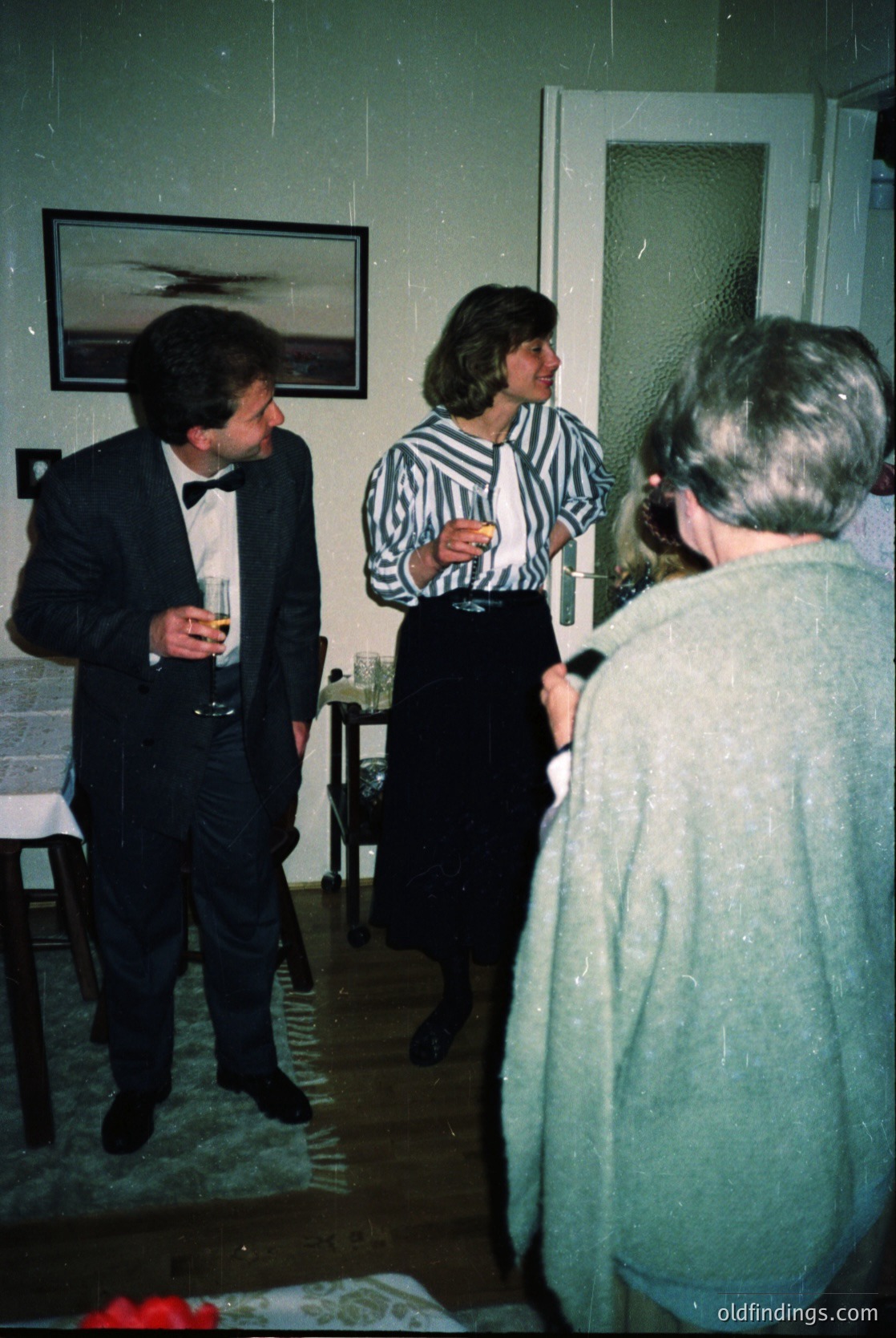 Formal indoor gathering in a mid-century interior, featuring three adults in 1960s-70s attire: a man in a dark suit with a bow tie, a woman in a striped blouse and pencil skirt, and another woman in a light sweater. The room includes a framed abstract art print, a small side table, and a doorway. Warm lighting and vintage color tones enhance the nostalgic ambiance.
