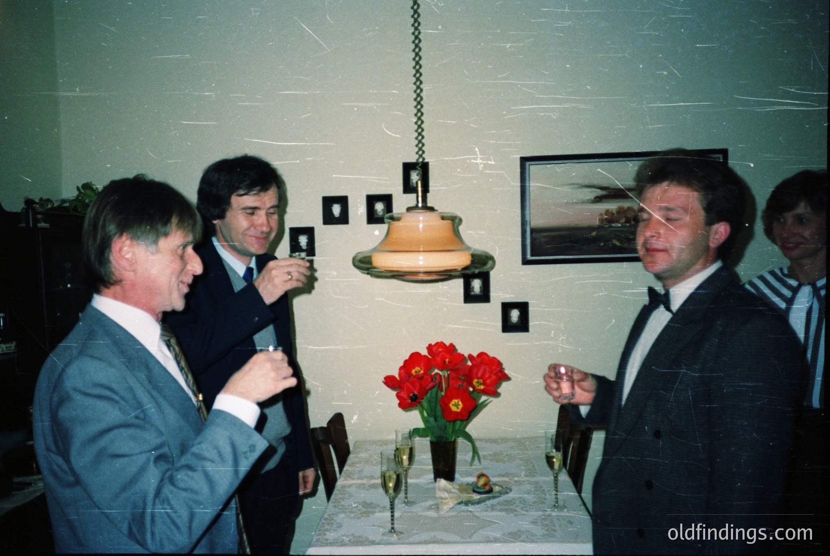 Four men in formal attire—dark suits, ties, and dress shirts—engage in conversation indoors, likely at a 1980s–1990s event. A vintage pendant light hangs above a table adorned with red tulips, champagne flutes, and a framed landscape print. Plain walls and minimal decor suggest a mid-century European setting.