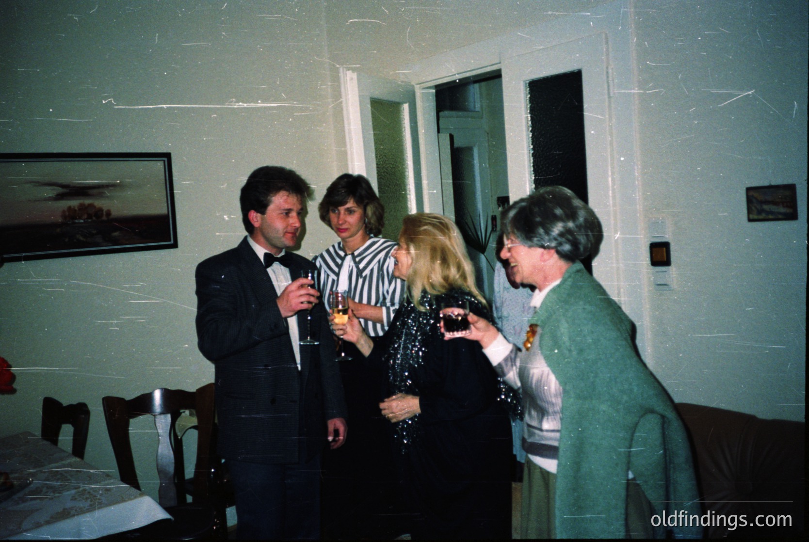 Four adults in formal attire gather indoors, likely at a 1980s–1990s event. Men wear suits with bow ties; women in blazers and dresses. One holds a champagne flute; another adjusts a decorative brooch. Plain walls, framed artwork, and wooden furniture suggest a modest venue. Potential corporate or celebratory occasion.