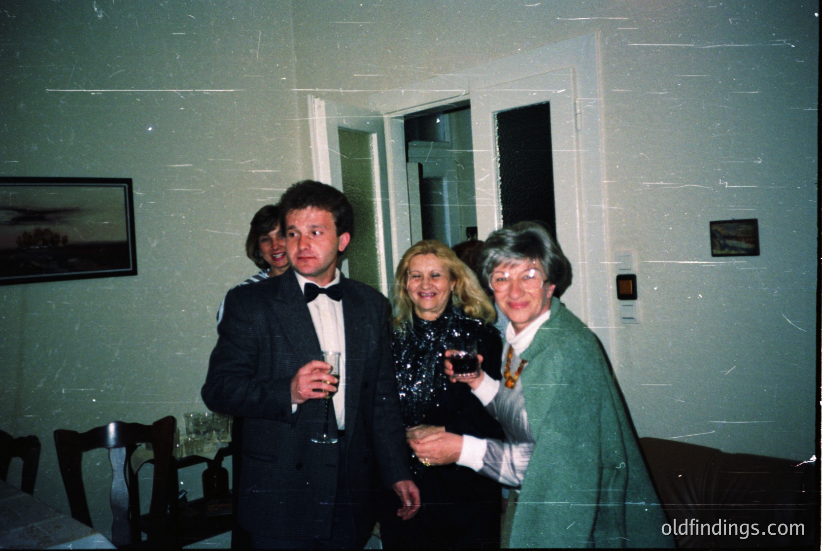 Three adults pose indoors, likely at a formal event in the 1990s. The man in a dark suit holds a wineglass; two women in formal attire (one in a green blazer, the other in a white top) hold drinks. Plain walls, framed art, and a door suggest a modest office or event space.