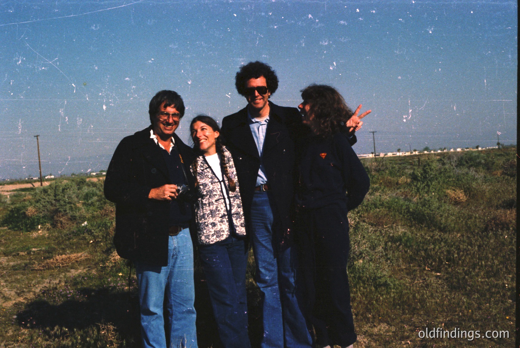 Four individuals pose outdoors in 1970s-style attire—men in blazers/sunglasses, women in patterned blouses and jeans—against a blurred industrial backdrop. Likely a group photo from the 1970s, possibly for a festival or event.