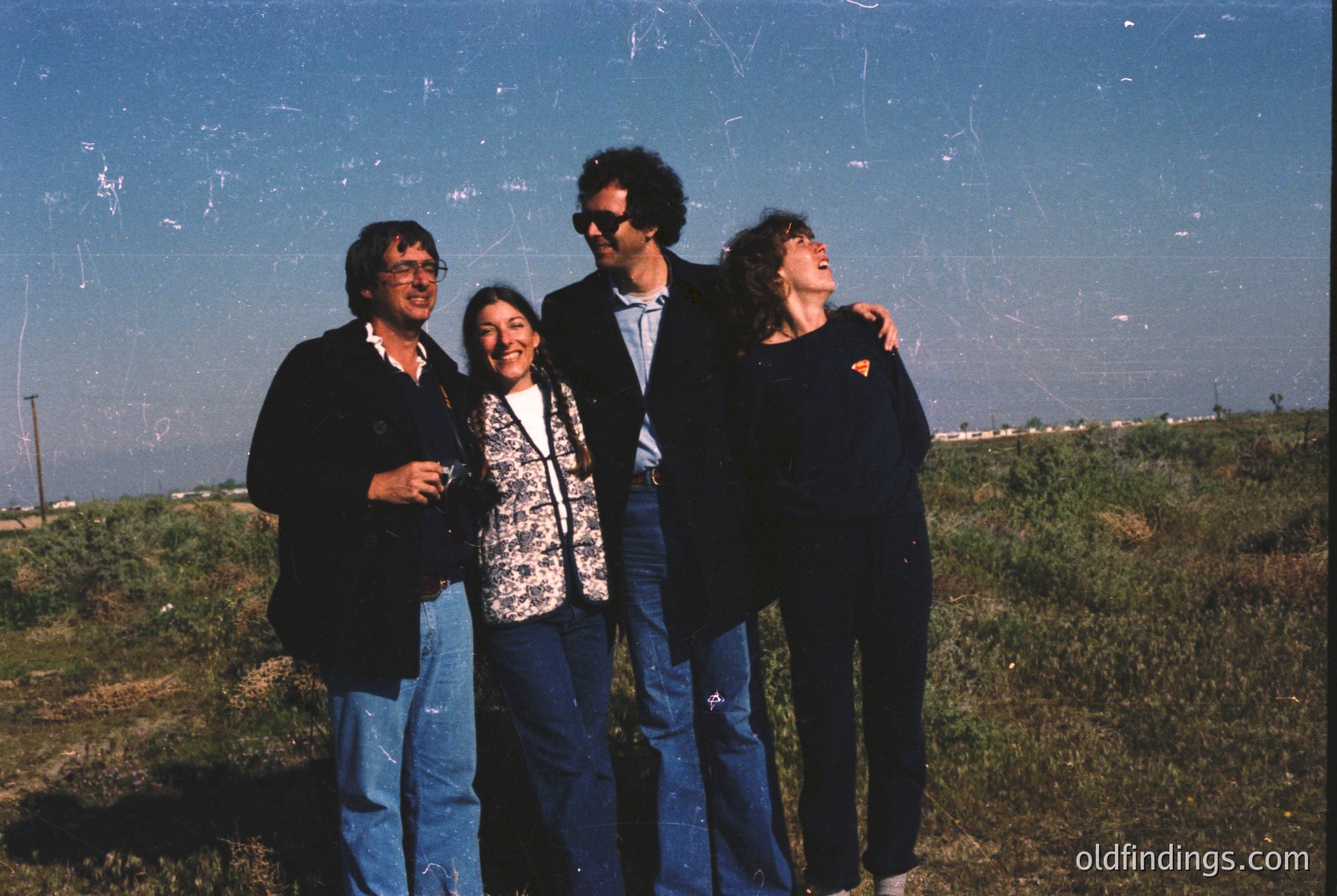 Four individuals pose outdoors in casual 1970s attire—men in blazers, women in patterned blouses and jeans—against a faded blue backdrop. Open grassy field with distant industrial structures suggests a rural or suburban setting. Authentic vintage aesthetic ideal for historical research or nostalgic design references.