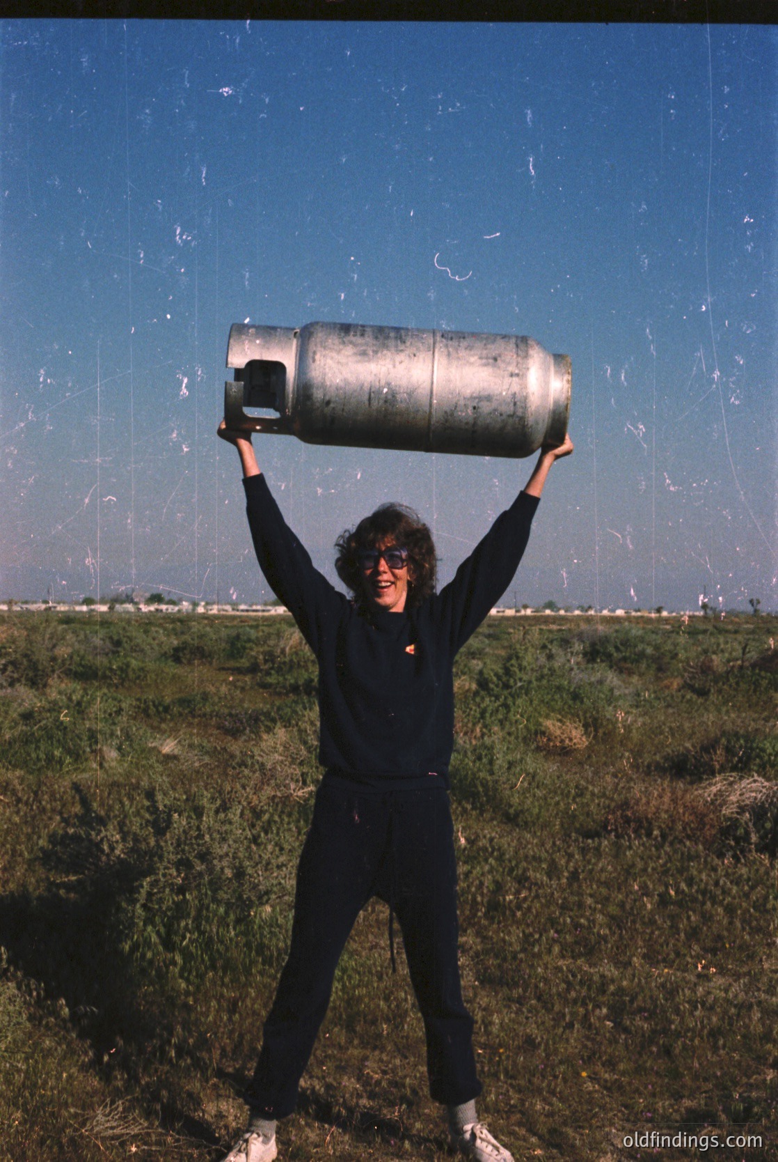 Person in outdoor setting holding large propane tank above head, likely mid-20th century. Clothing suggests casual, utilitarian style—sweater, pants, and sneakers. Background shows industrial or agricultural landscape with flat terrain and clear sky.
