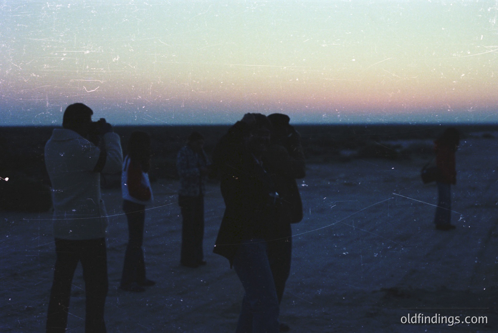 Silhouetted group of photographers at dusk, silhouetted against a soft sunset horizon. Vintage film grain overlay enhances nostalgic, artistic texture. Coastal or open landscape with minimal visible detail.