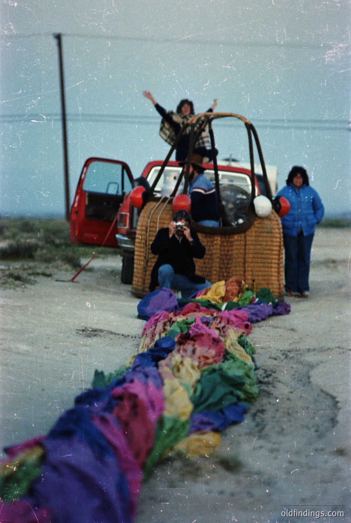 Vintage photo of a lively outdoor scene featuring a red vintage car with wicker luggage. A person stands atop the trunk, arms raised, while another kneels beside a colorful, folded blanket or textile display. A third person in winter attire stands nearby. Likely 1960s–1970s, possibly a roadside market or festival.