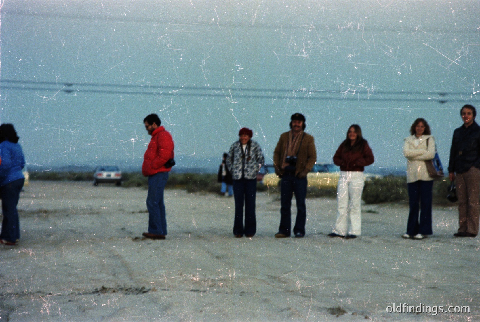 Vintage group photo on a concrete beachfront, likely 1970s–1980s. Six individuals in casual 70s attire—plaid shirts, bell-bottoms, jackets—pose near a large, smooth concrete wall. One person holds a camera, suggesting a leisure trip. Coastal setting with minimal vegetation and a distant horizon.