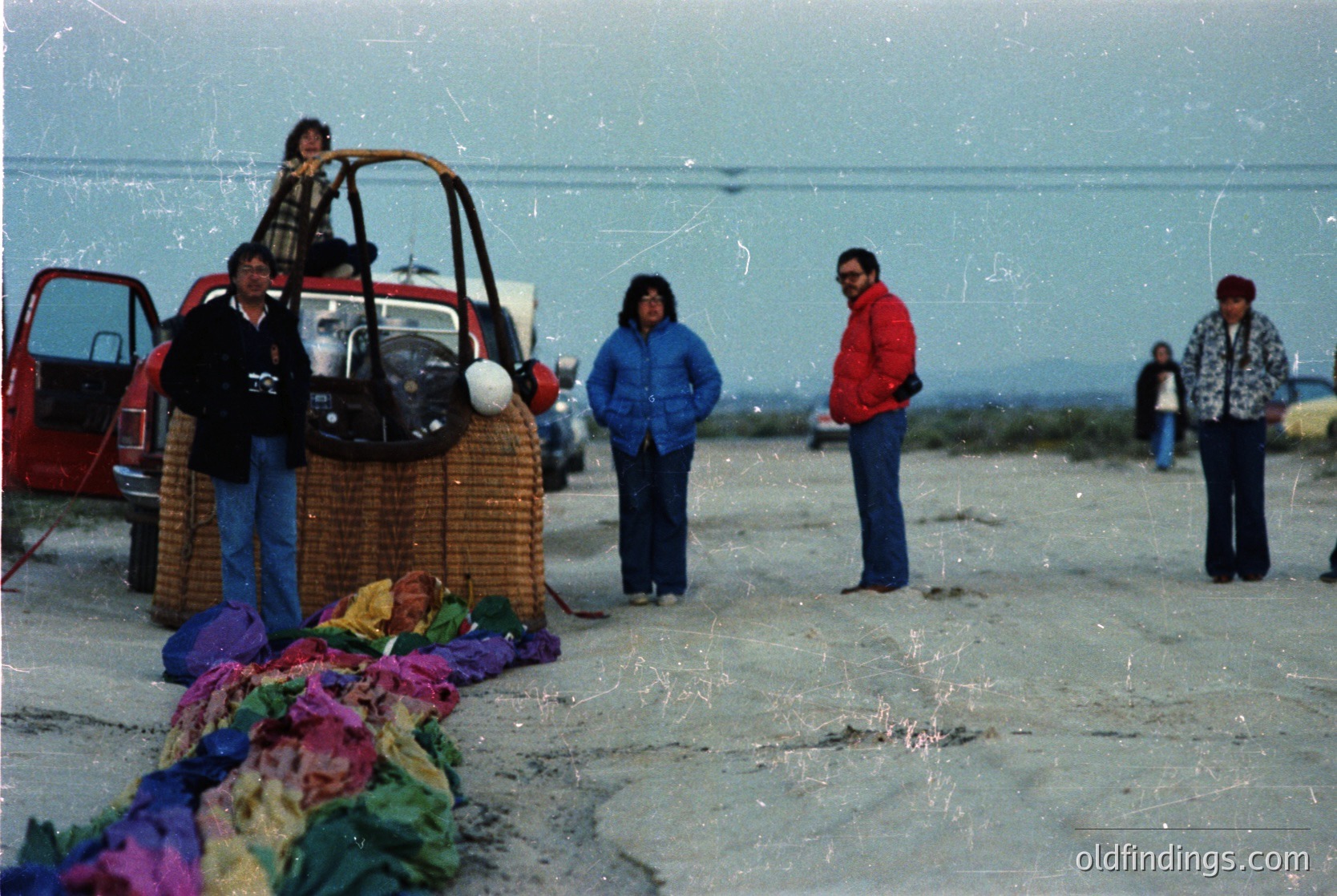 Vintage seaside scene with group posing near a vintage Fiat 500. Colorful woven baskets and textiles laid on pavement, suggesting a market or gathering. Overcast skies and coastal backdrop hint at a Northern European or Atlantic location. Mid-20th century attire and car indicate or .