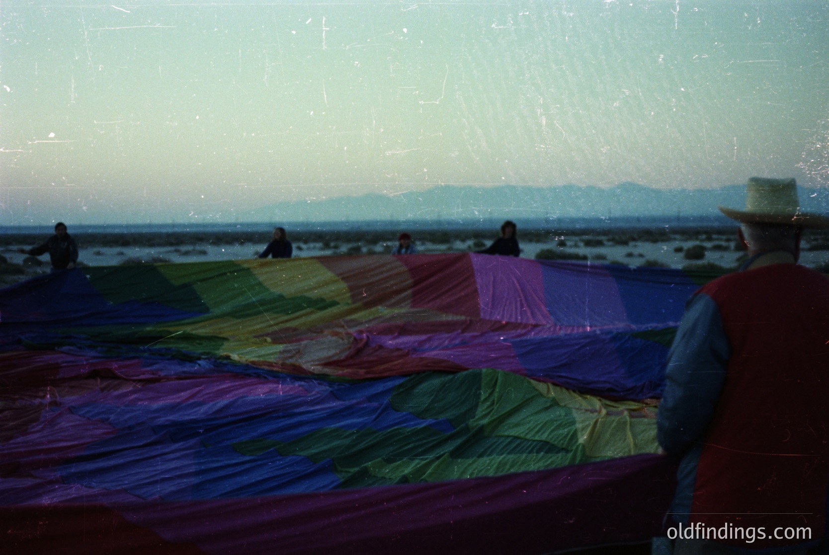 Vintage seaside scene featuring vibrant, hand-dyed fabric drying on a long rack. Four figures—two in foreground, two in background—appear engaged in drying or arranging textiles. Coastal horizon with distant mountains under soft, hazy light. Likely Mediterranean or Adriatic region, 1960s–1980s.