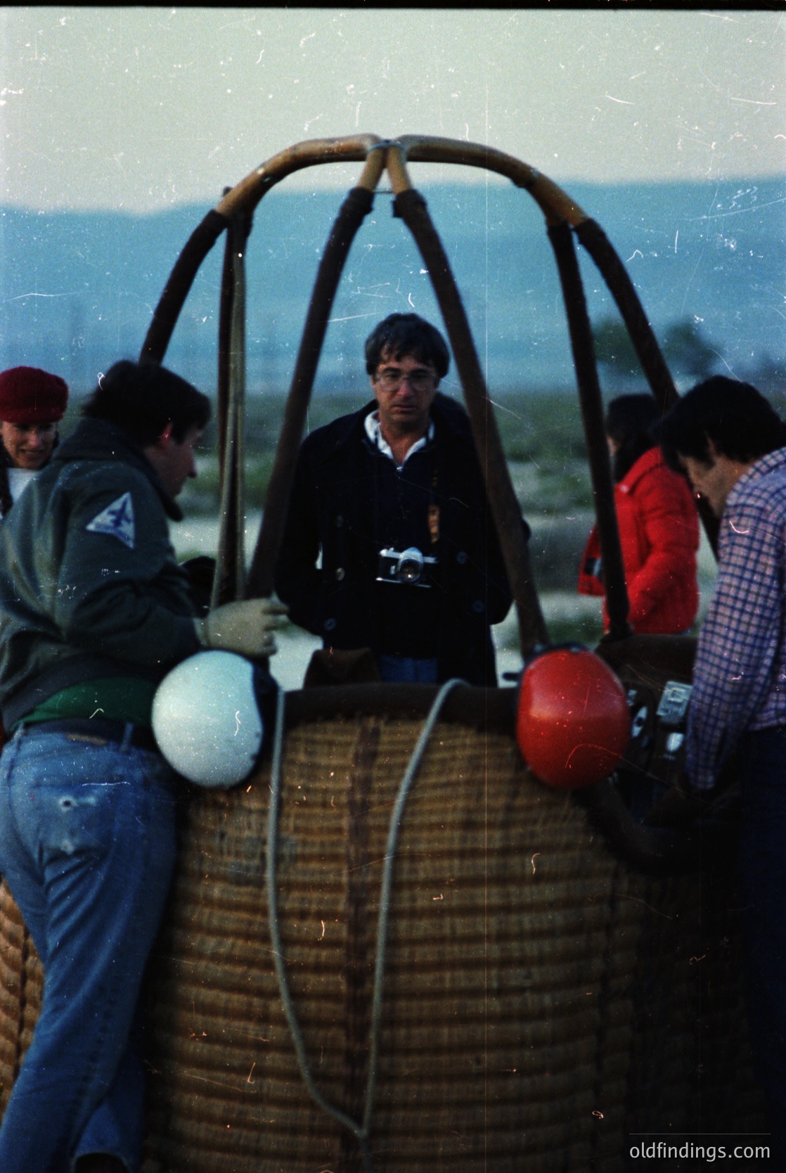 Hot-air balloon basket with woven rattan frame, carrying four individuals in casual 1970s attire—one holding a camera, another in a red jacket. Open landscape with distant mountains and flat terrain suggests a rural or desert setting. Vintage grain and sepia tone indicate archival quality.