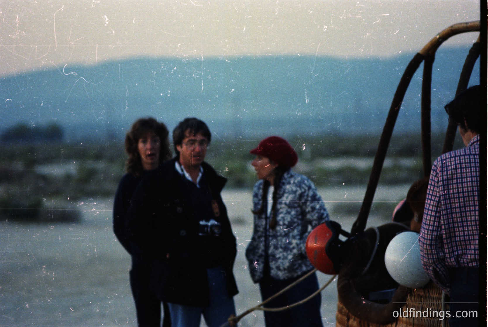 Vintage photo of four individuals on a boat, likely a ferry or fishing vessel, with mountainous coastal backdrop. The scene suggests a 1970s–1980s European seaside location. Distinctive elements include a life preserver, rope handles, and a basket. The faded film grain and clothing styles hint at mid-century maritime culture.