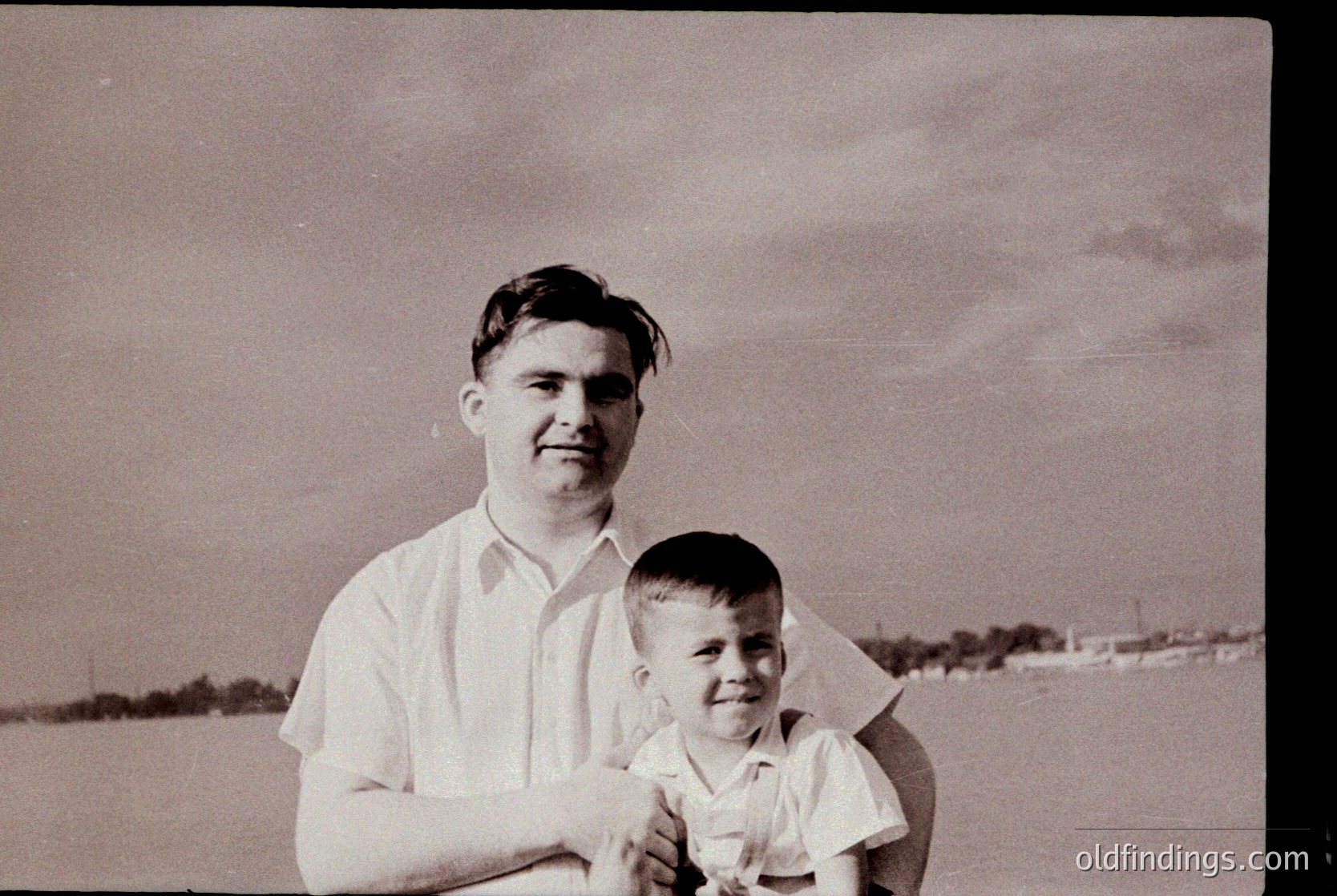 Black-and-white portrait of a man and young boy in outdoor setting, likely mid-20th century. Man wears short-sleeved dress shirt; boy in short-sleeved button-up with rolled cuffs. Background shows flat terrain with distant fence and trees, suggesting rural or suburban area.
