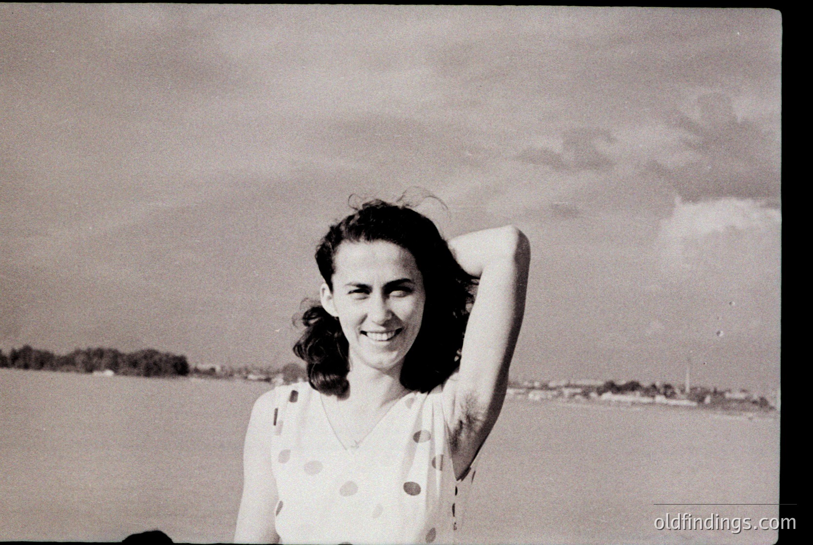 Vintage black-and-white portrait of a woman in a polka-dot dress, hand on head, smiling against a blurred seaside background. Mid-20th century beachwear and candid expression evoke 1950s–1960s coastal leisure.