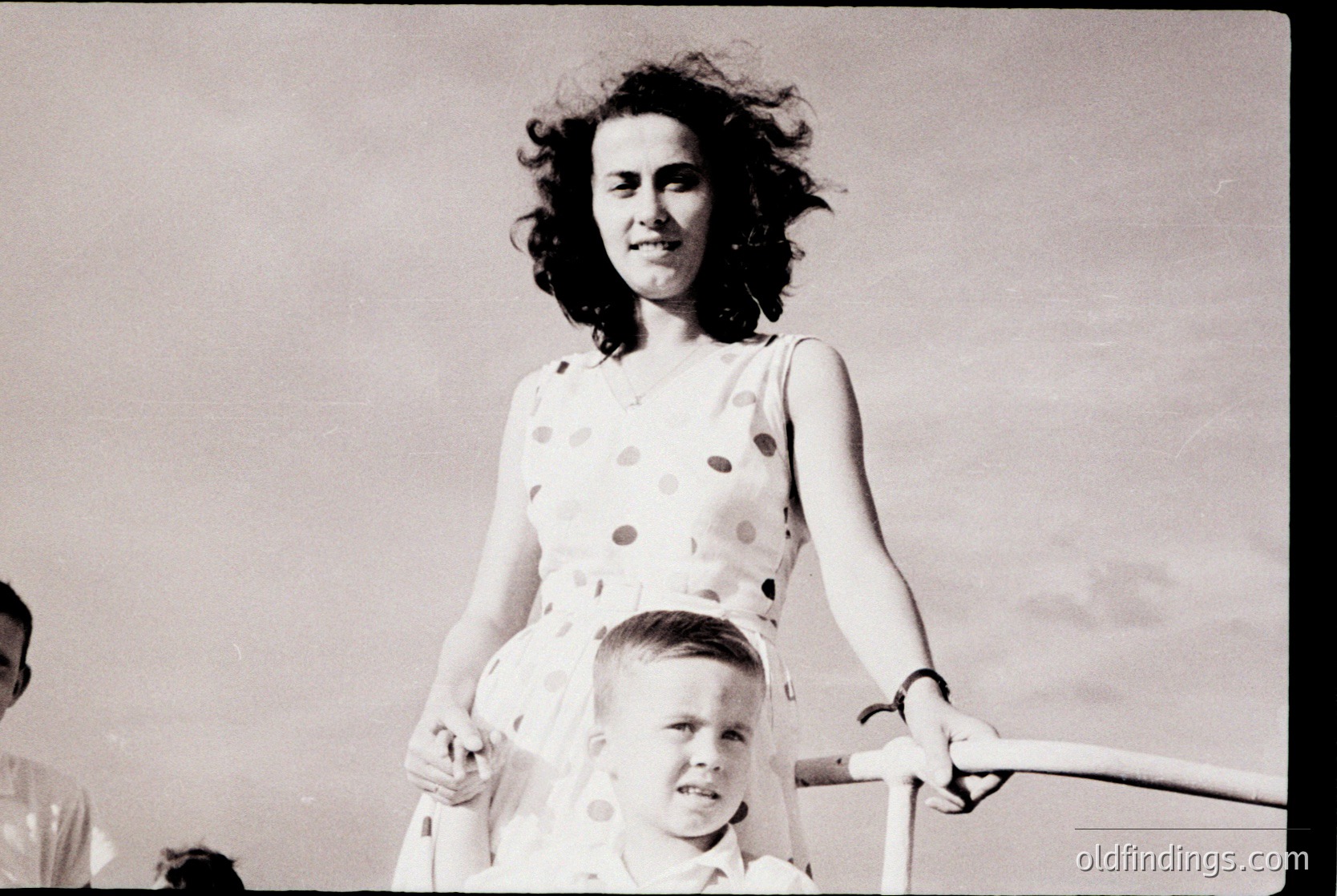 Black-and-white candid of a woman in a polka-dot dress (1950s–60s style) holding a young boy on a carousel horse, mid-ride. Crowd and blurred motion suggest lively outdoor amusement park setting.