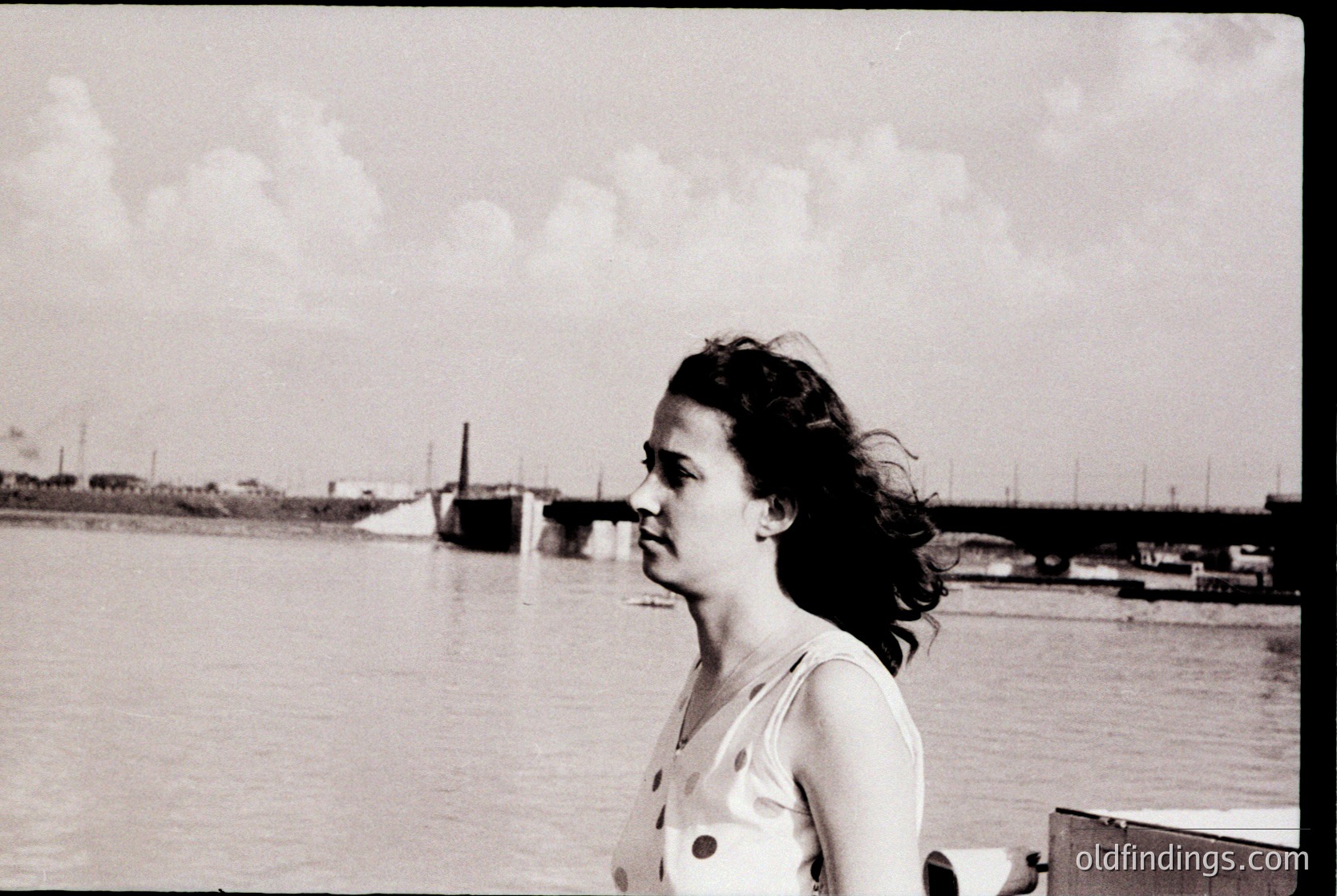 Black-and-white portrait of a woman in mid-20th century attire—polka-dotted blouse—standing near a waterfront with industrial docks and a bridge in background. Soft focus and candid composition evoke mid-century documentary style. Likely or .