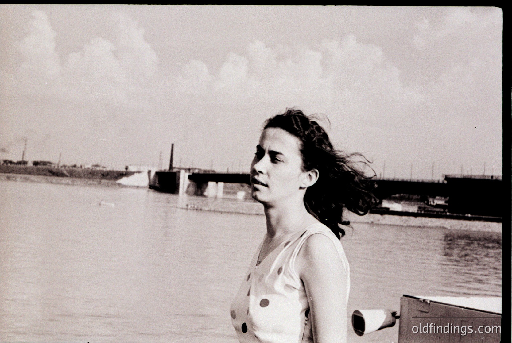 Black-and-white portrait of a woman in a sleeveless dress with button details, standing near a waterfront with industrial docks and a bridge in the background. Mid-20th century fashion and urban coastal setting.