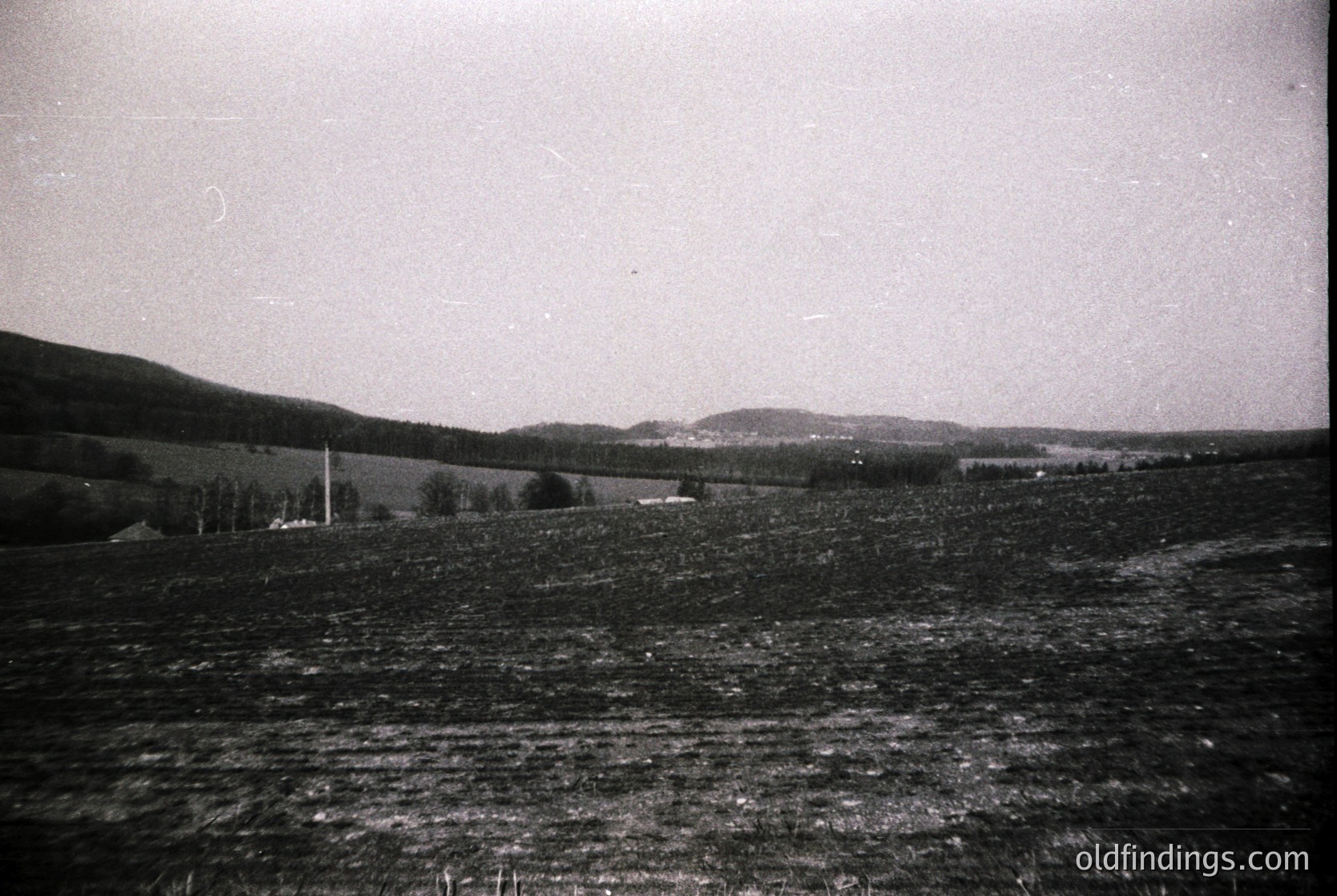 Vintage black-and-white landscape featuring rolling hills and sparse farmland. A single isolated house and scattered trees mark the horizon. Grainy texture suggests mid-20th century photography. Ideal for historical or nostalgic design references.