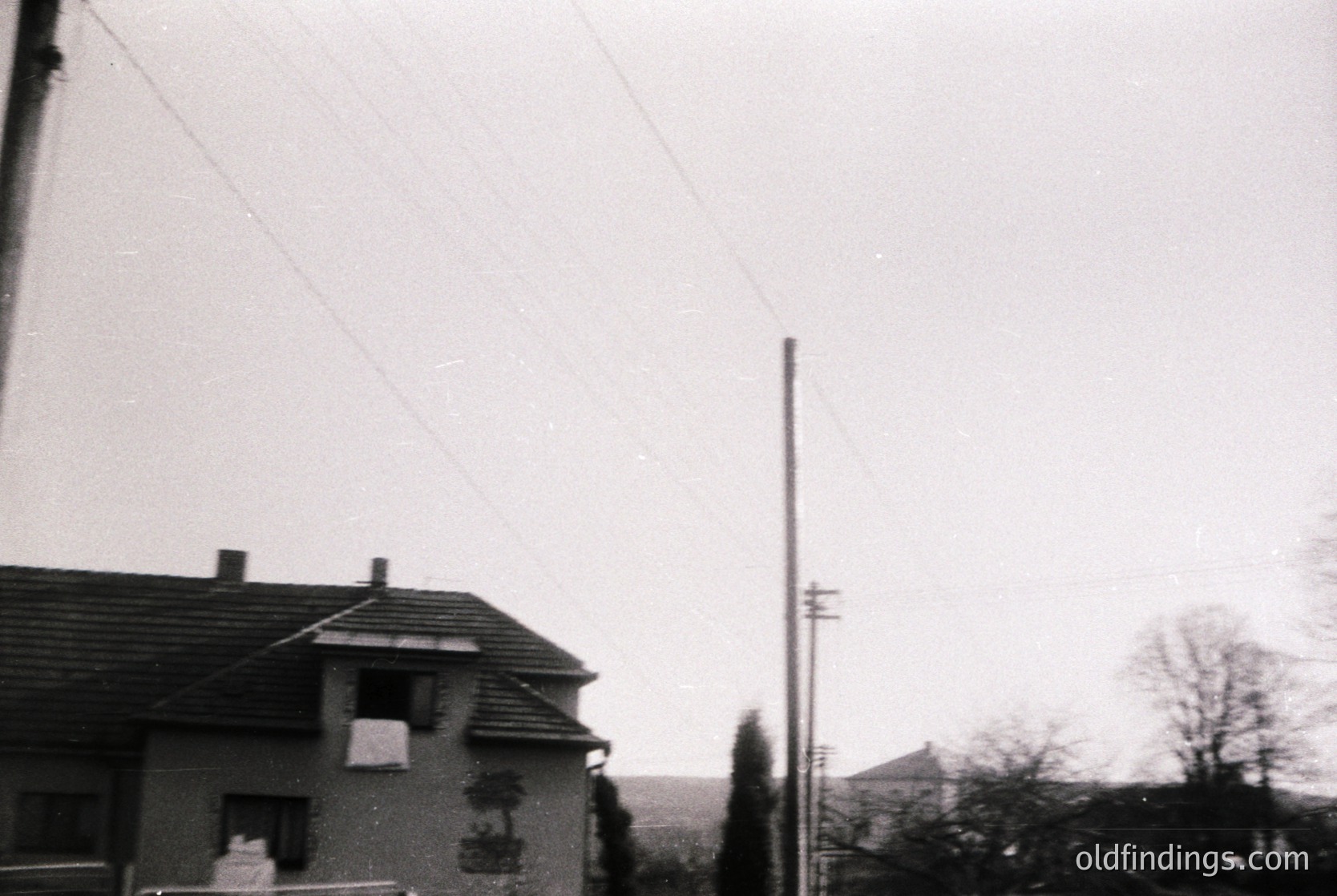 Vintage black-and-white shot of a modest residential building with a tiled roof and chimney, set against a muted landscape. Overhead power lines and utility poles frame the scene, suggesting mid-20th-century urban/suburban architecture. Minimalist design with a coat of arms or emblem on the facade.