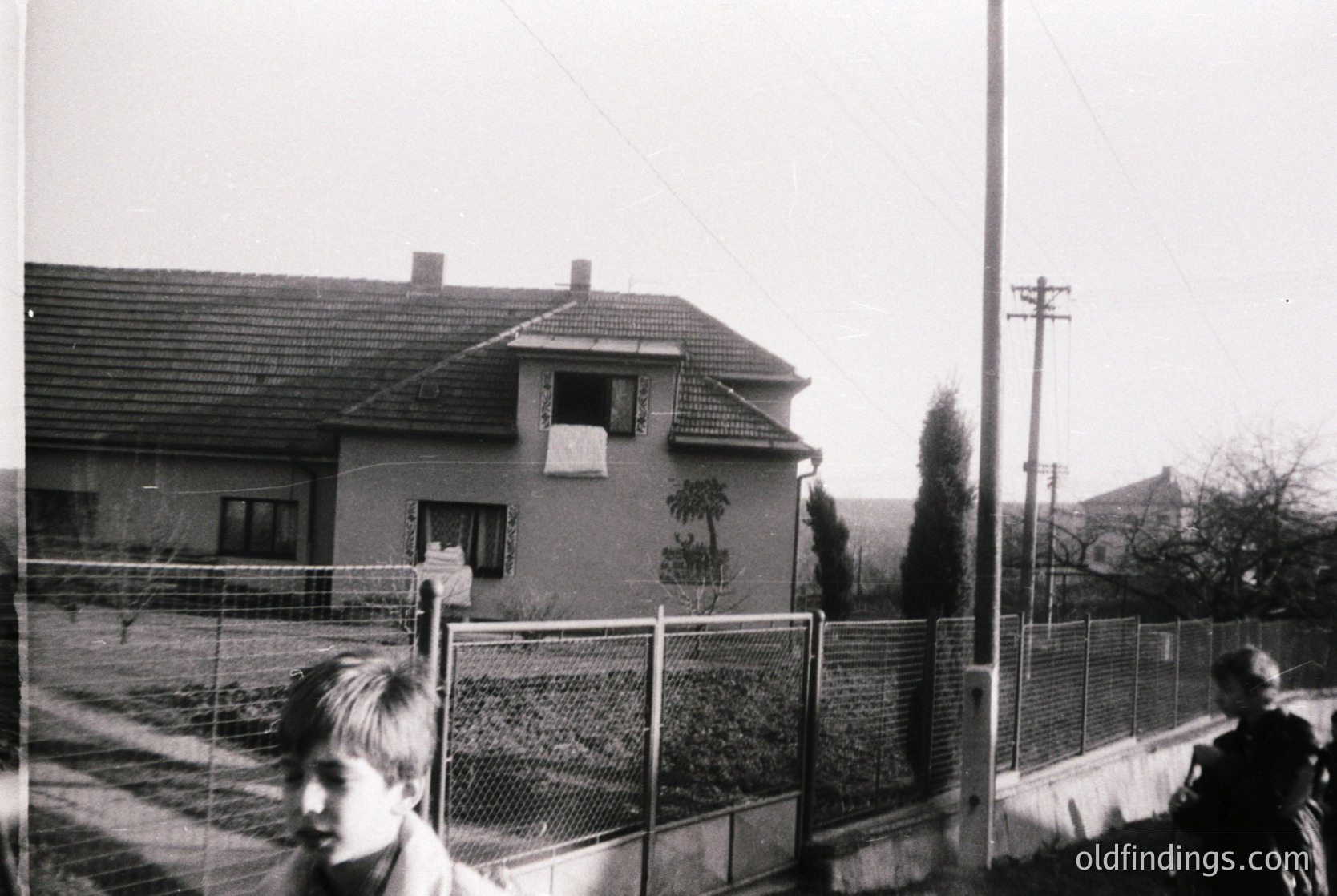 Mid-20th century residential street with Soviet-era brick house featuring mural of palm tree. Fenced yard and barbed wire atop fence suggest restricted access. Two children in foreground, one blurred. Overhead power lines and sparse trees indicate urban/suburban setting. Likely Eastern Bloc region, 1960s–1980s.
