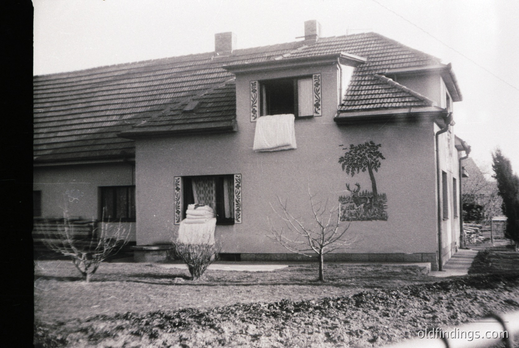 Two-story residential building with decorative plasterwork, likely Eastern European . Red-tiled roof, small front yard with young trees, and a mural depicting rural scenery. Functional, modest suburban design.