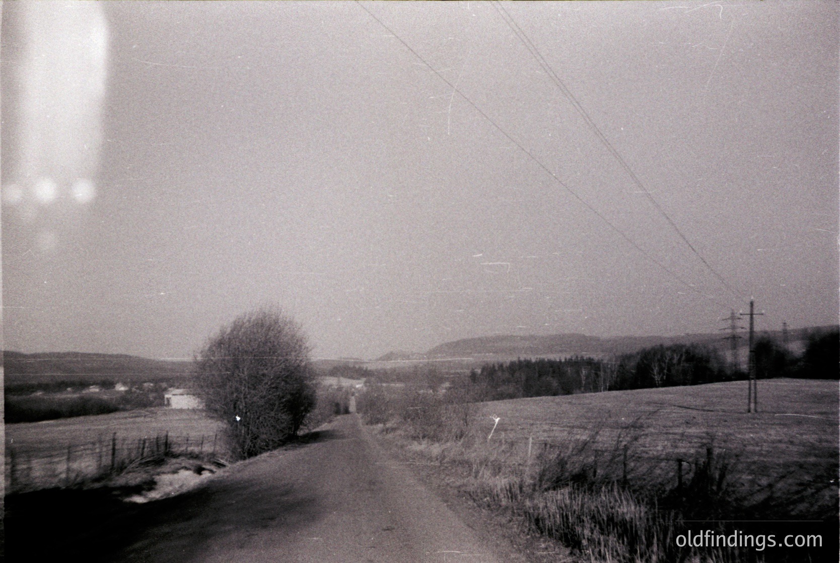 Vintage black-and-white rural road winding through open fields, bordered by sparse trees and utility poles. Overcast sky and distant hills suggest early-to-mid 20th century European countryside. Faded film grain and slight vignette enhance nostalgic atmosphere.