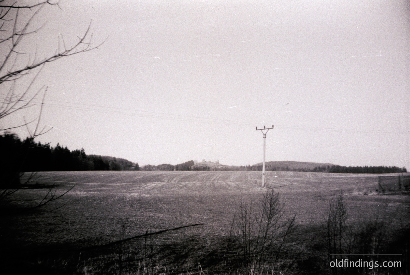 Vintage black-and-white rural landscape featuring plowed fields under an overcast sky. A lone communication tower stands in the center distance, surrounded by sparse trees and barren branches. Mid-20th century agricultural setting, likely Eastern Europe.