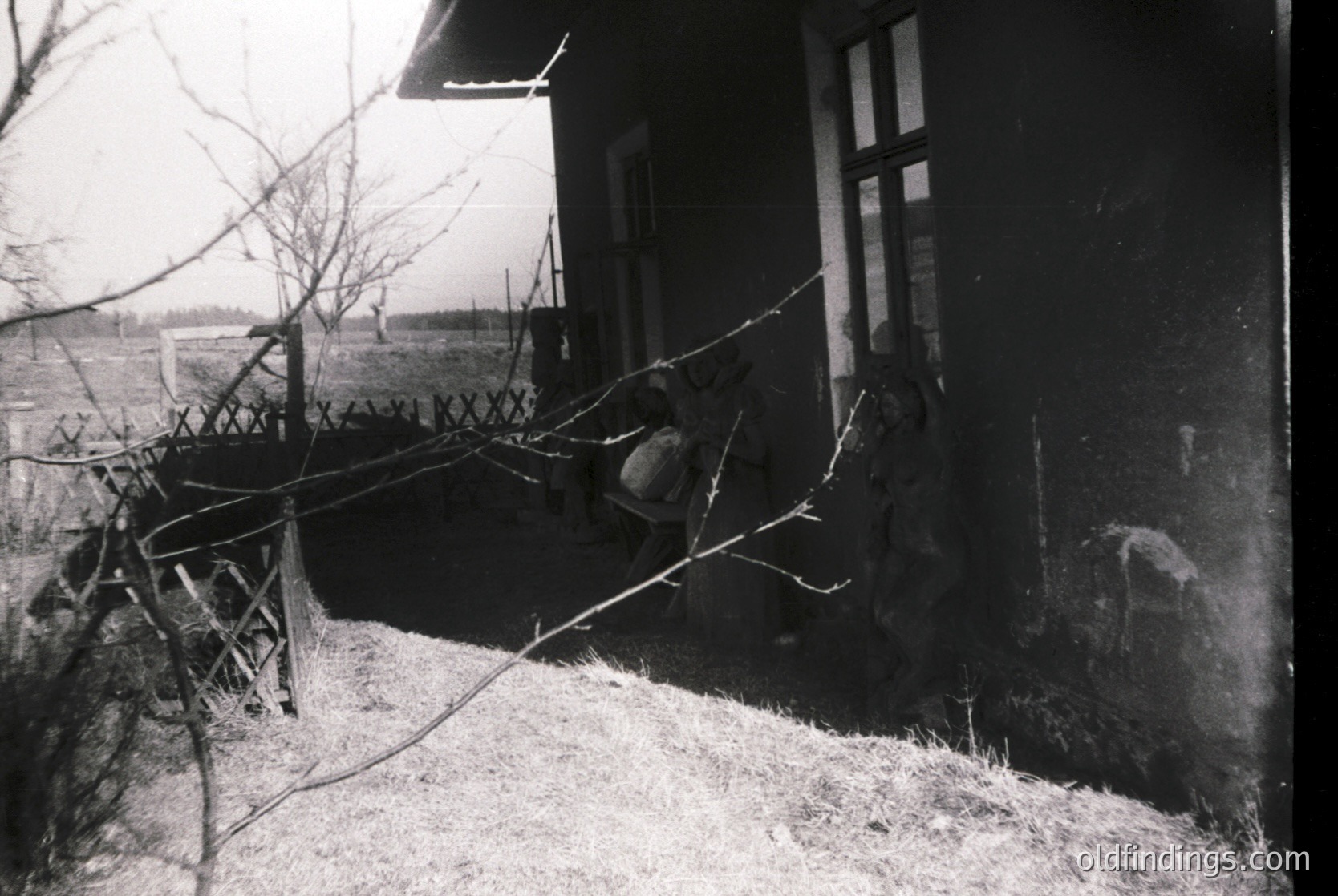 Vintage black-and-white photo of a rural porch with a child sitting on a makeshift swing made from branches. Wooden house with peeling paint, barbed wire fence, and leafless trees. Likely Eastern European countryside, mid-20th century.