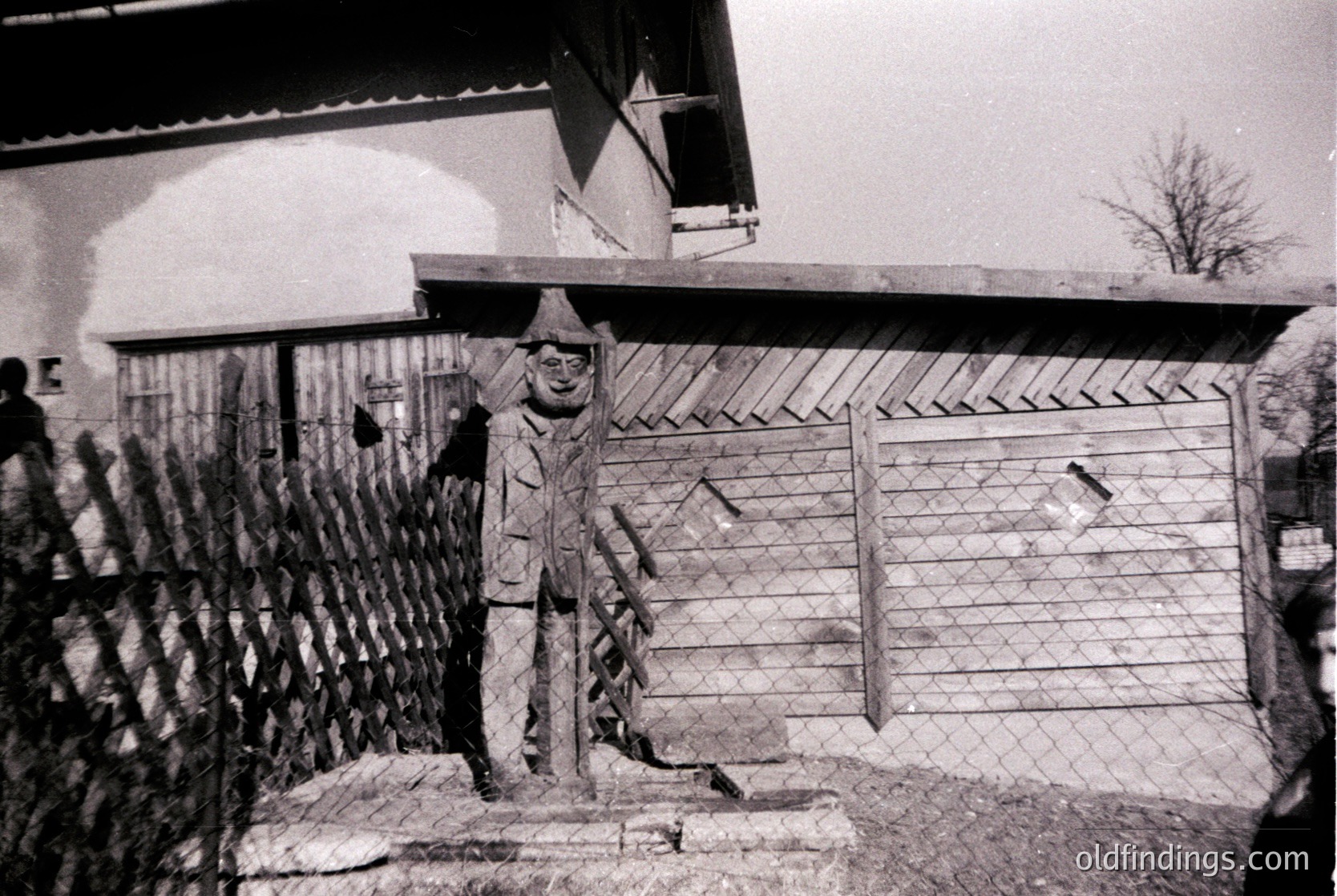 Black-and-white portrait of a man in a winter coat, hat, and gloves standing beside a rustic brick chimney and corrugated fence. Mid-20th century rural European setting.