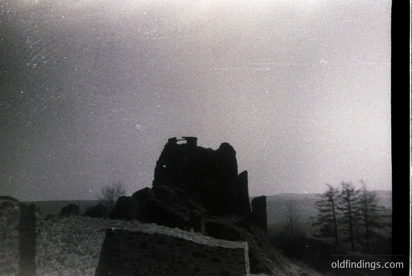 Mid-century black-and-white shot of a ruined stone tower atop rocky terrain, silhouetted against a starry night sky. Minimalist composition highlights architectural decay and celestial contrast. Likely –1960s European landscape photography.