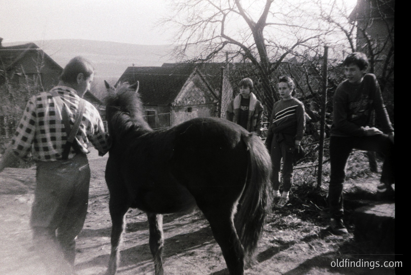 Black-and-white rural scene featuring a man in a plaid shirt leading a horse along a dirt path, with three onlookers—two women and a man—standing nearby. Wooden houses and leafless trees suggest late autumn/winter. Mid-20th century European countryside setting.