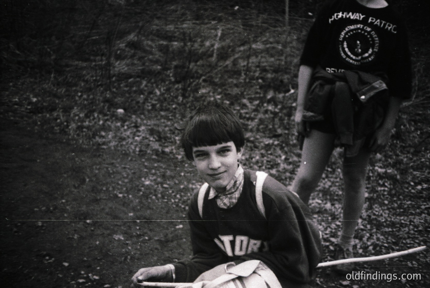 Black-and-white photo of a young boy in a vintage "Toronto" sports jersey, seated outdoors on gravel. Partial view of another figure in a "Highway Patrol" uniform in background. Mid-20th century urban setting, likely 1950s–1960s.