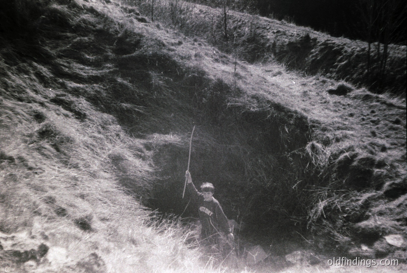 A lone figure in mid-stride casts a long shadow across rugged terrain, framed by sparse vegetation. The individual appears to be hiking or climbing, holding a long pole or staff. The scene suggests a high-altitude or alpine environment, likely from the mid-20th century based on the monochrome and clothing style. [Mid-20th century alpine hiker with staff, rugged mountain terrain, high-contrast black & white ]