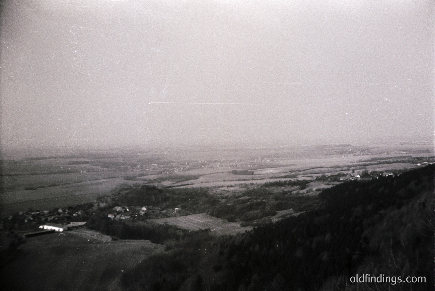 Vintage aerial view of a rural landscape with scattered farmhouses and fields, likely mid-20th century. Low-lying hills and dense tree clusters frame the horizon.