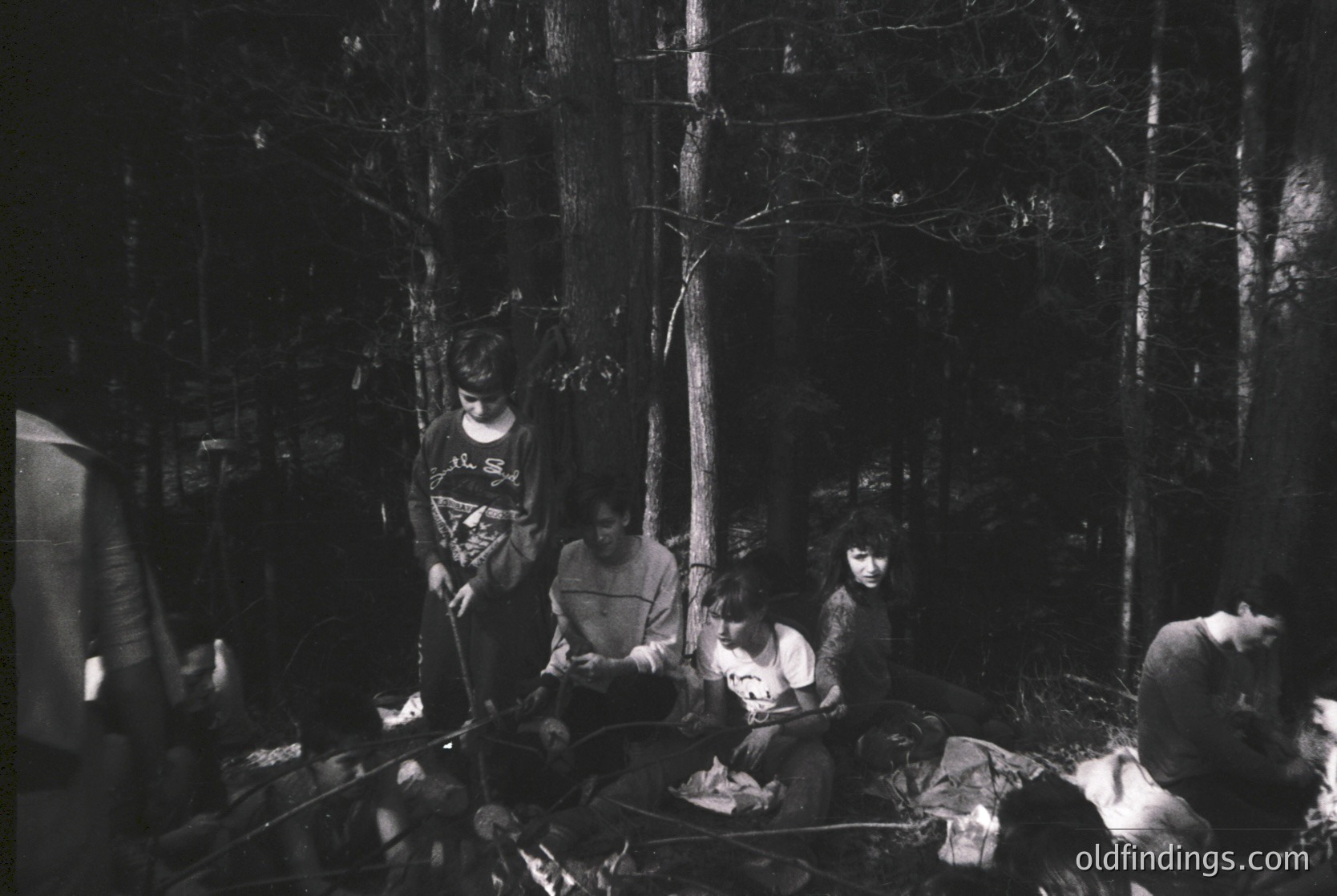 Four individuals gathered around a campfire in a dense forest, seated on logs and blankets. Mid-20th century outdoor lifestyle captured in monochrome.
