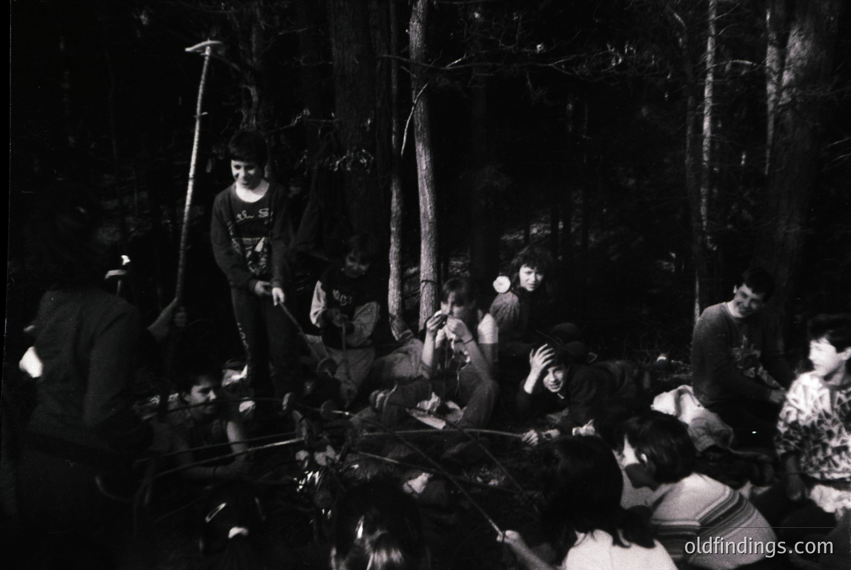 Black-and-white campfire scene in dense forest, likely 1970s. Group of 10+ people gathered around a fire pit, seated on logs. One person stands holding a long tool, possibly an axe. Casual, informal gathering suggests outdoor recreation or communal activity.