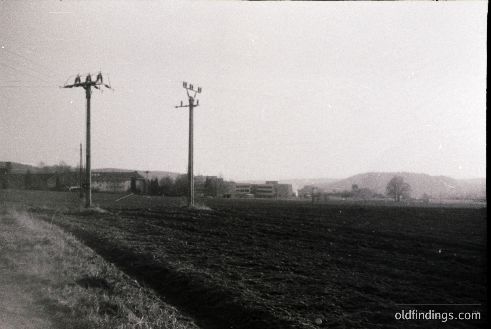 Mid-20th century rural landscape with two utility poles and barbed wire fence. Open fields stretch toward low-lying buildings and distant hills under overcast skies.